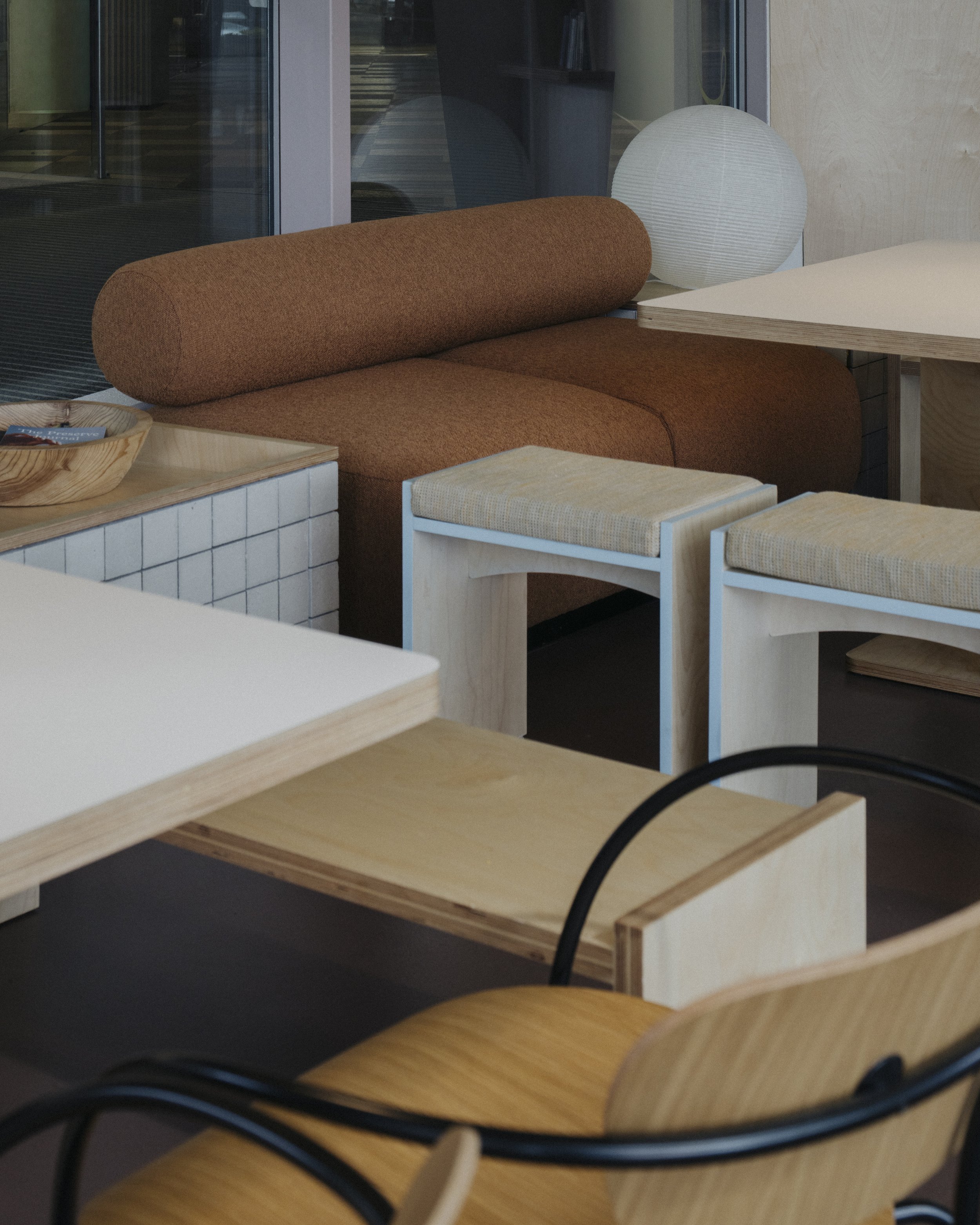 A cozy interior seating area with a brown fabric sofa, light-colored wooden tables and chairs, and a modern white paper lantern lamp.