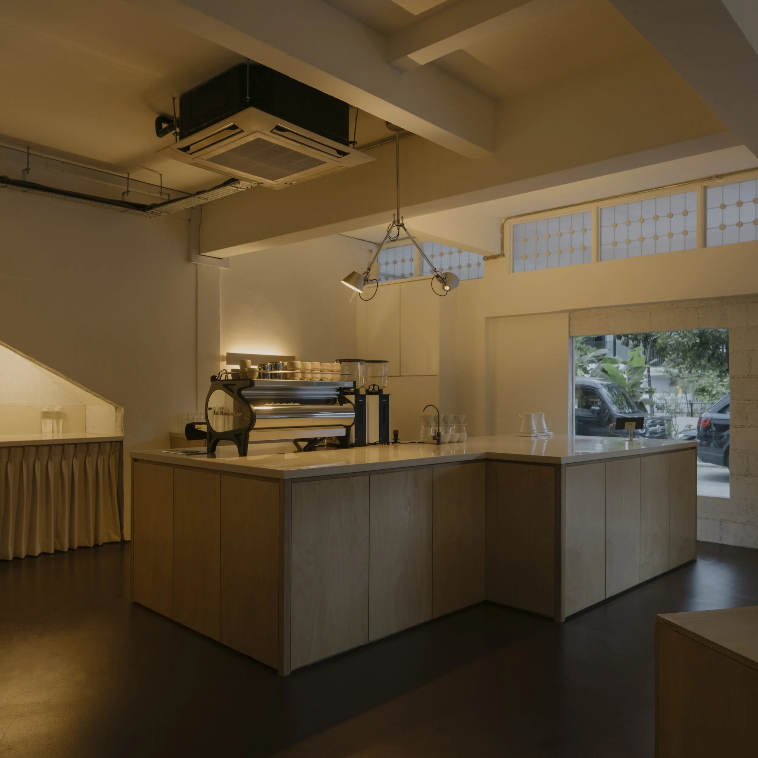 A minimalist coffee shop interior with a light wood counter, espresso machine, stacked cups, and large window with plants outside.