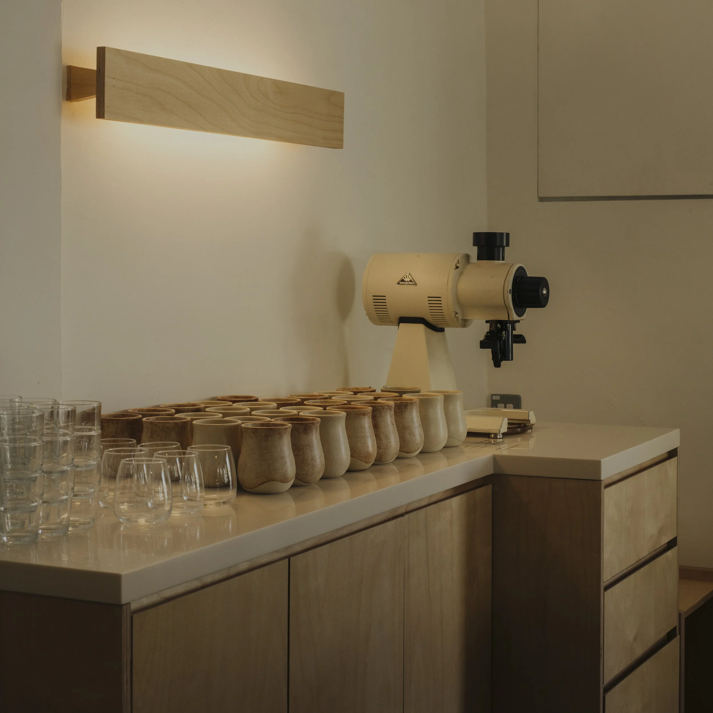 Wooden candle holders and glasses on a modern wooden cabinet, with a vintage beige microscope and a wall-mounted wooden light fixture in a minimalist room.