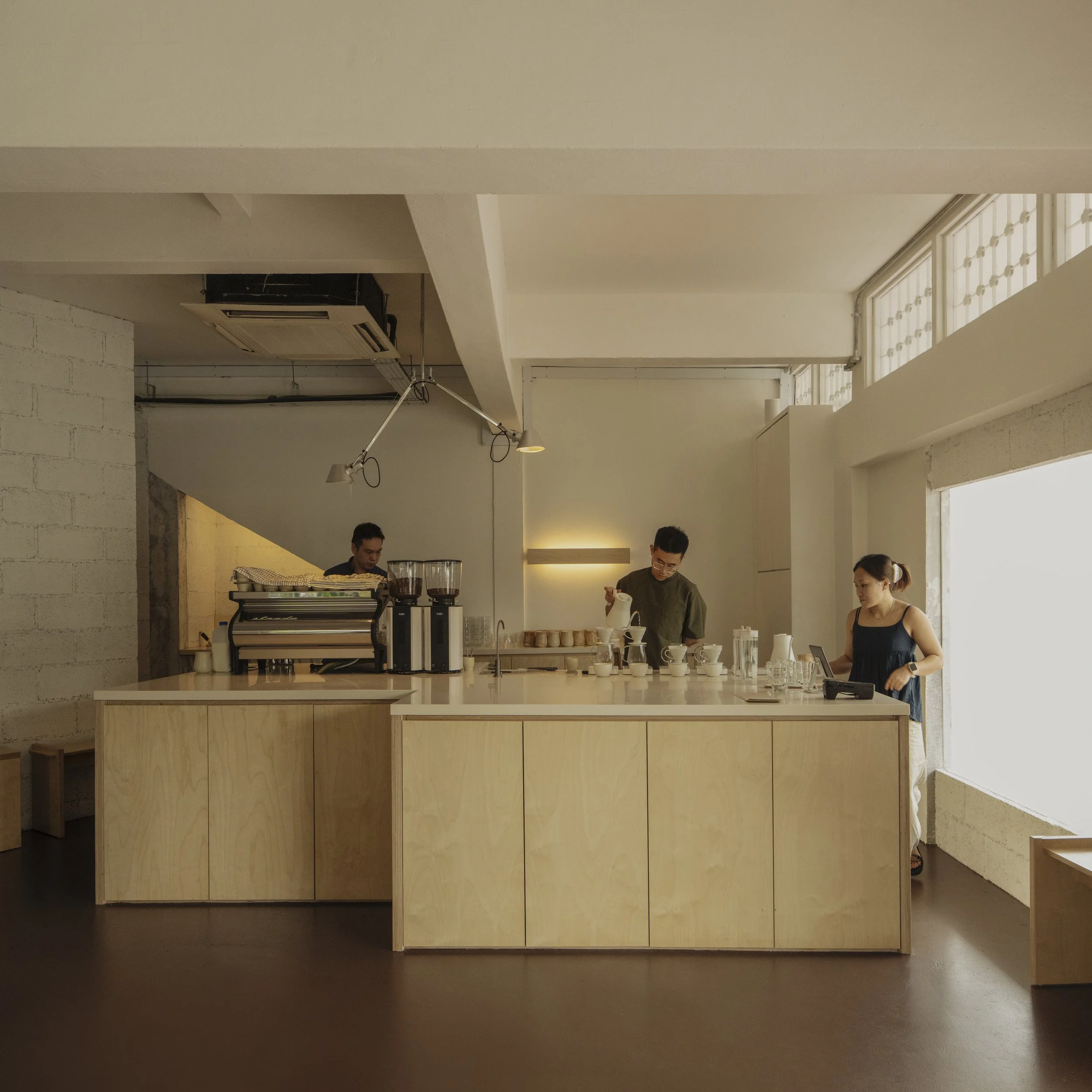 A minimalistic coffee shop interior with a large window, a wooden counter, and attention to natural light, featuring three staff members preparing drinks.