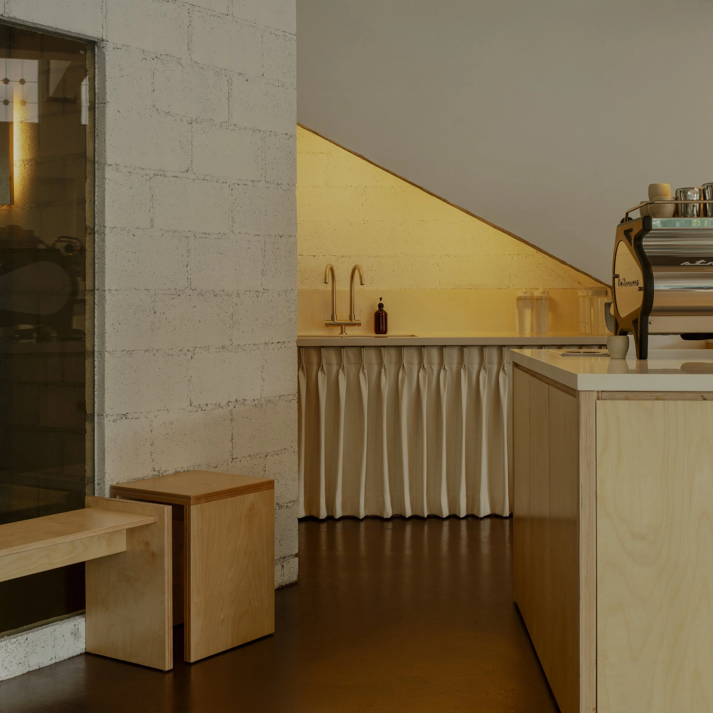 Interior view of a minimalist coffee shop with wooden furniture, a small sink with a gold faucet under a slanted ceiling, and a stainless steel espresso machine on a light wooden counter.