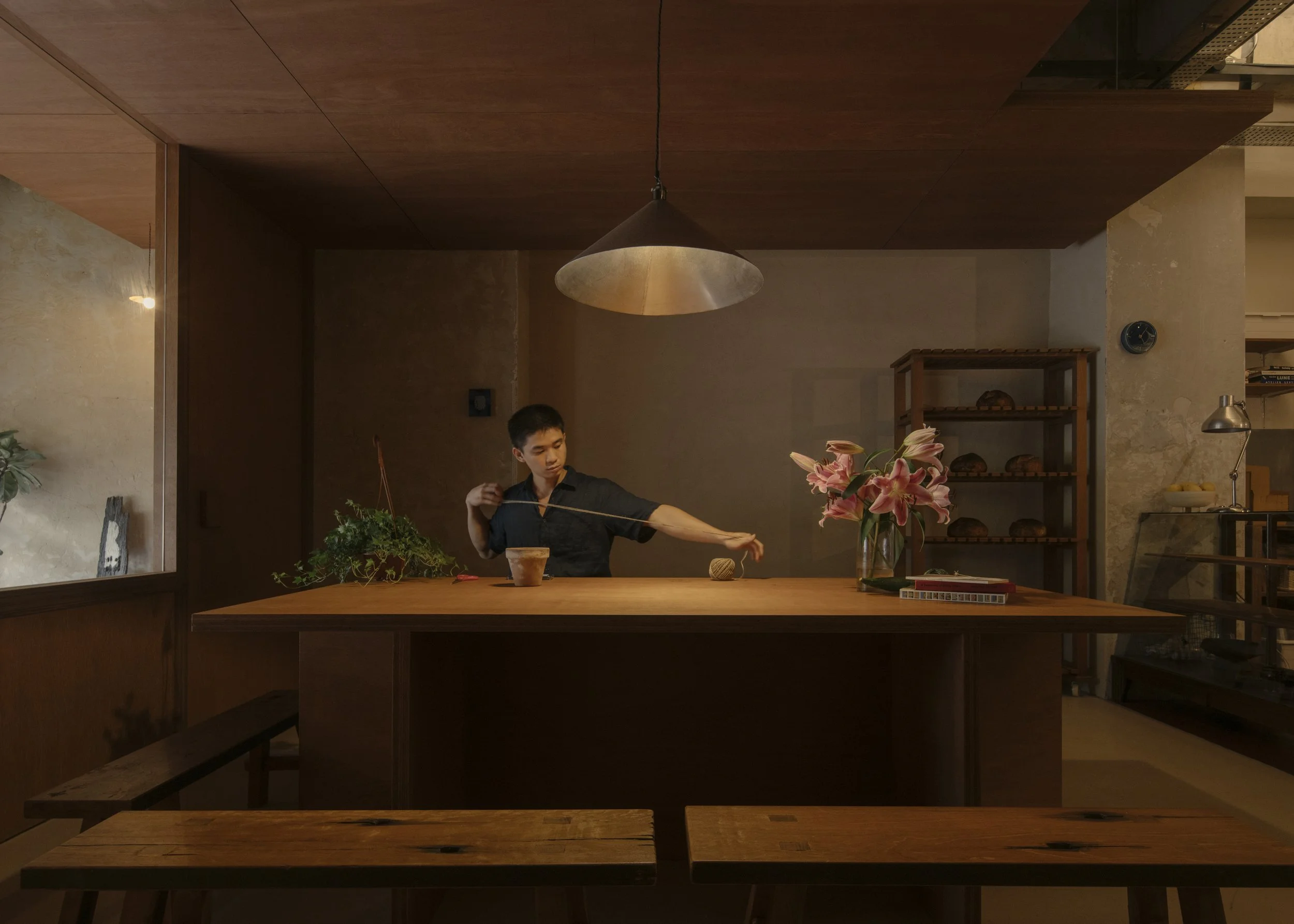A young man is sitting at a wooden dining table, doing floral arrangement in a nicely lit room decorated with flowers, plants, and shelves of bread.