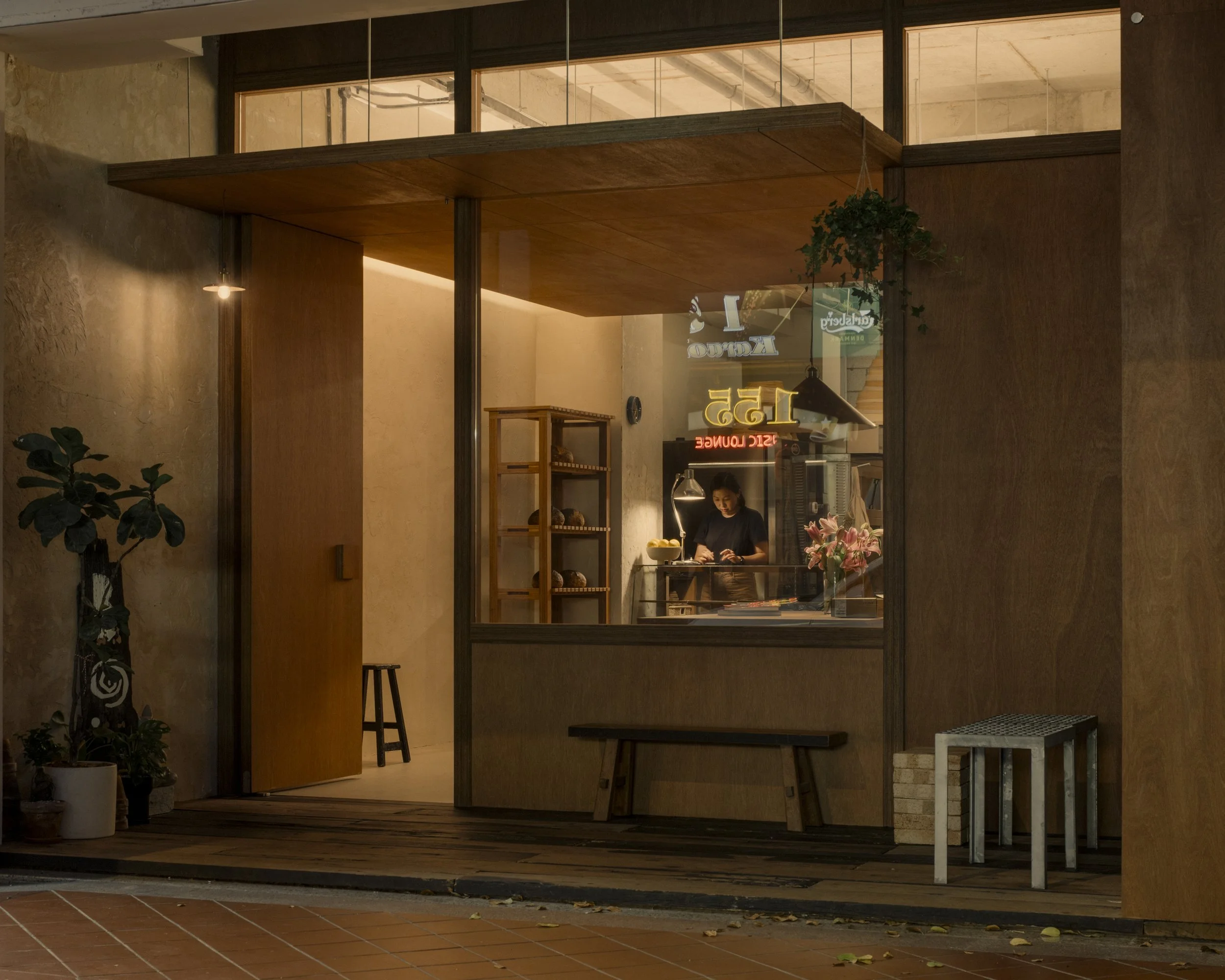 View into a cozy coffee shop or bakery with warm lighting, wooden furniture, and potted plants. A person is working behind the counter with a neon sign and various baked goods on shelves visible through the window.