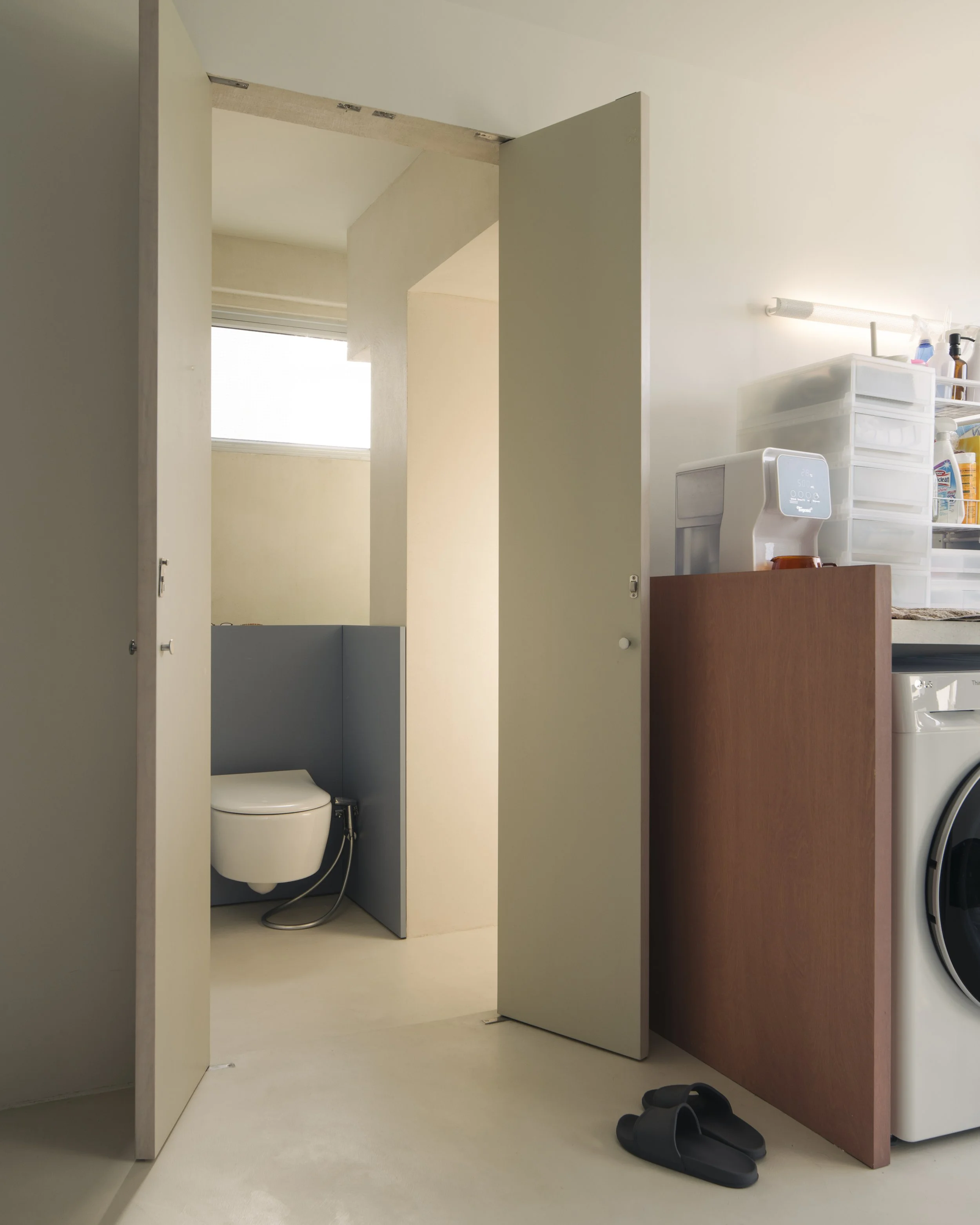 Open bathroom door revealing a toilet in a small space with a window above, next to a laundry area with shelves, storage drawers, a water dispenser, and a washing machine, with slippers on the floor.