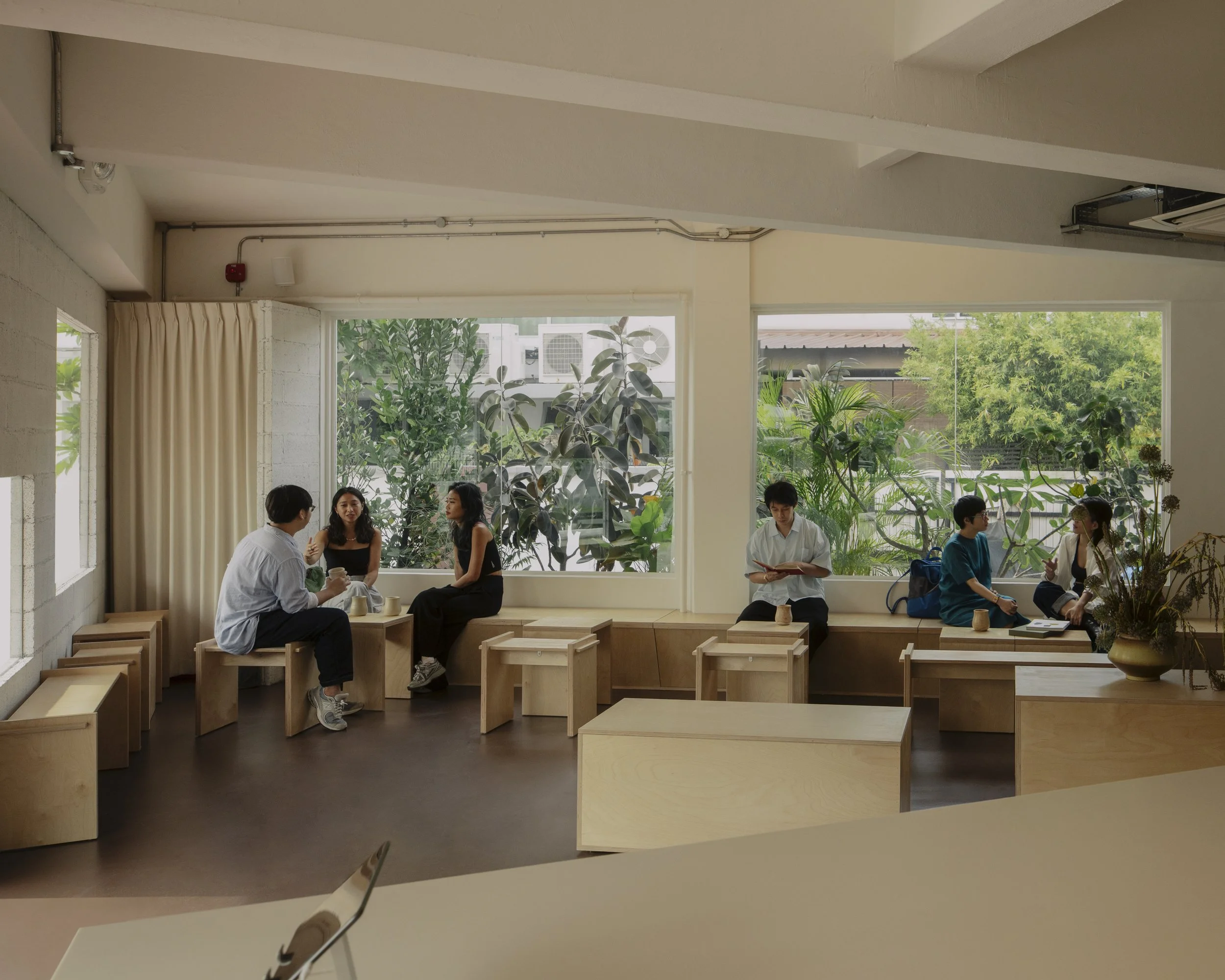 People sitting and talking inside a modern, well-lit room with large windows and greenery outside.
