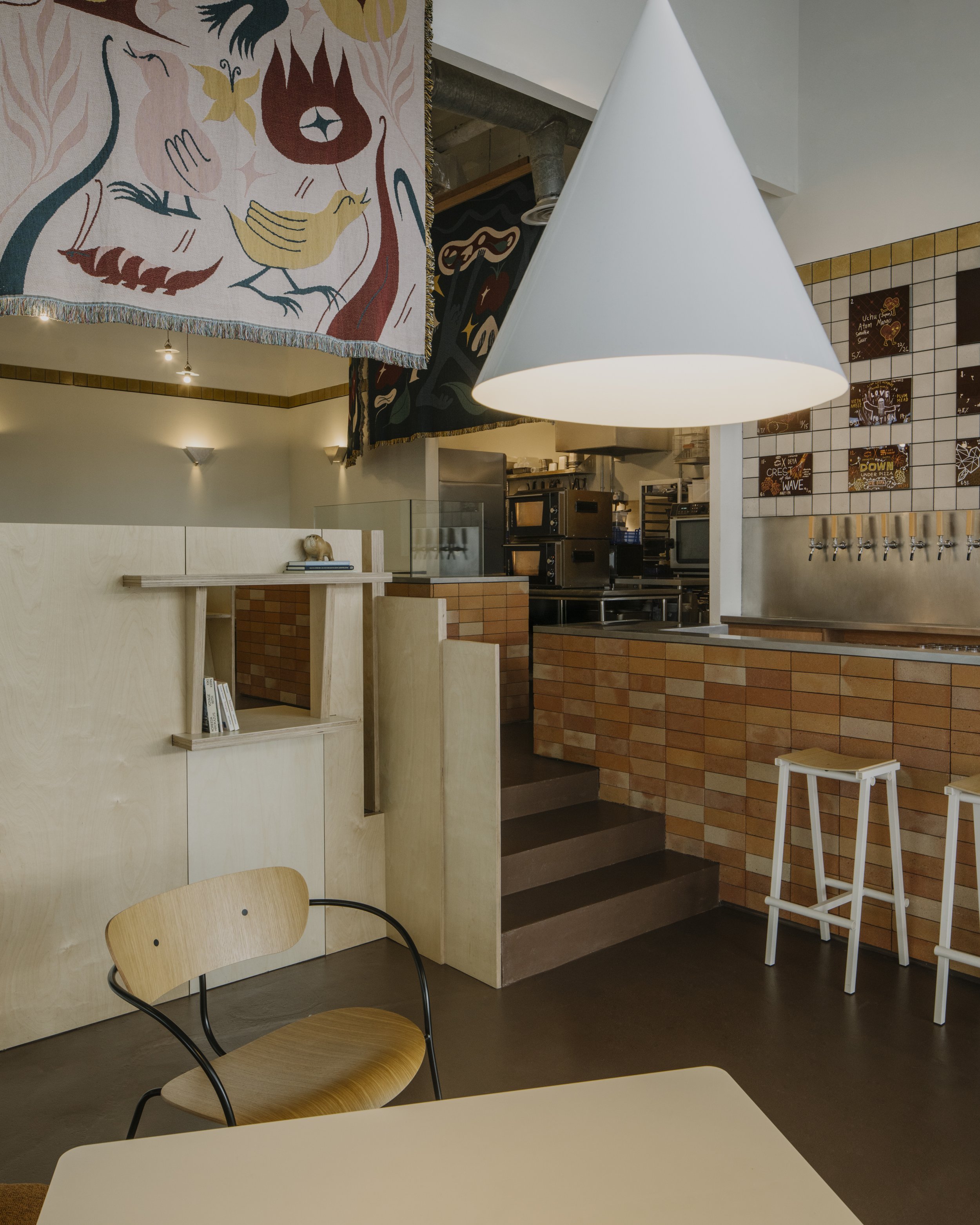 Interior of a modern cafe with wooden furniture, hanging art, a large white pendant light, and a brick counter with taps.