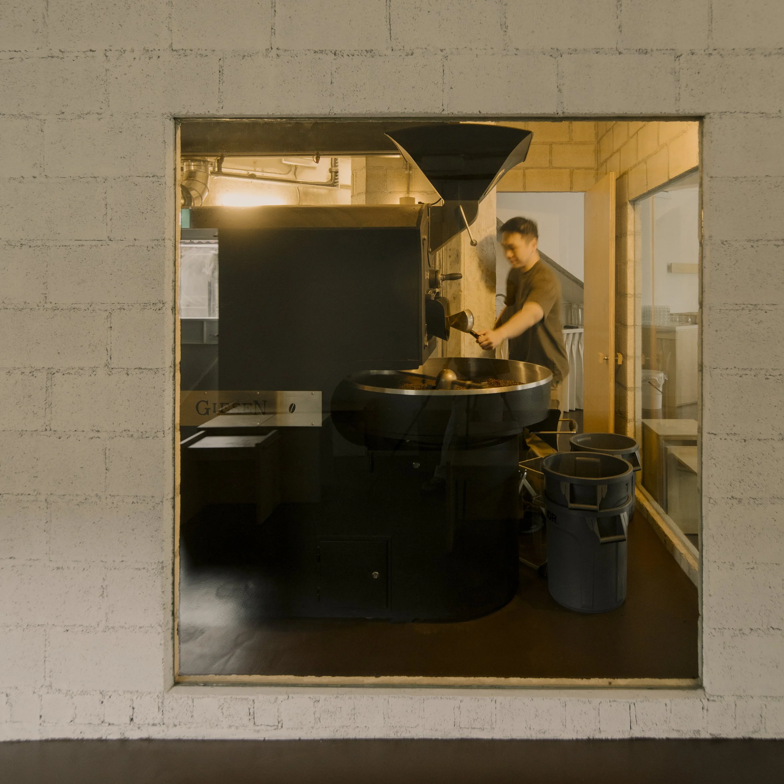 A person is operating a large coffee roasting machine in a room viewed through a square window in a white brick wall.