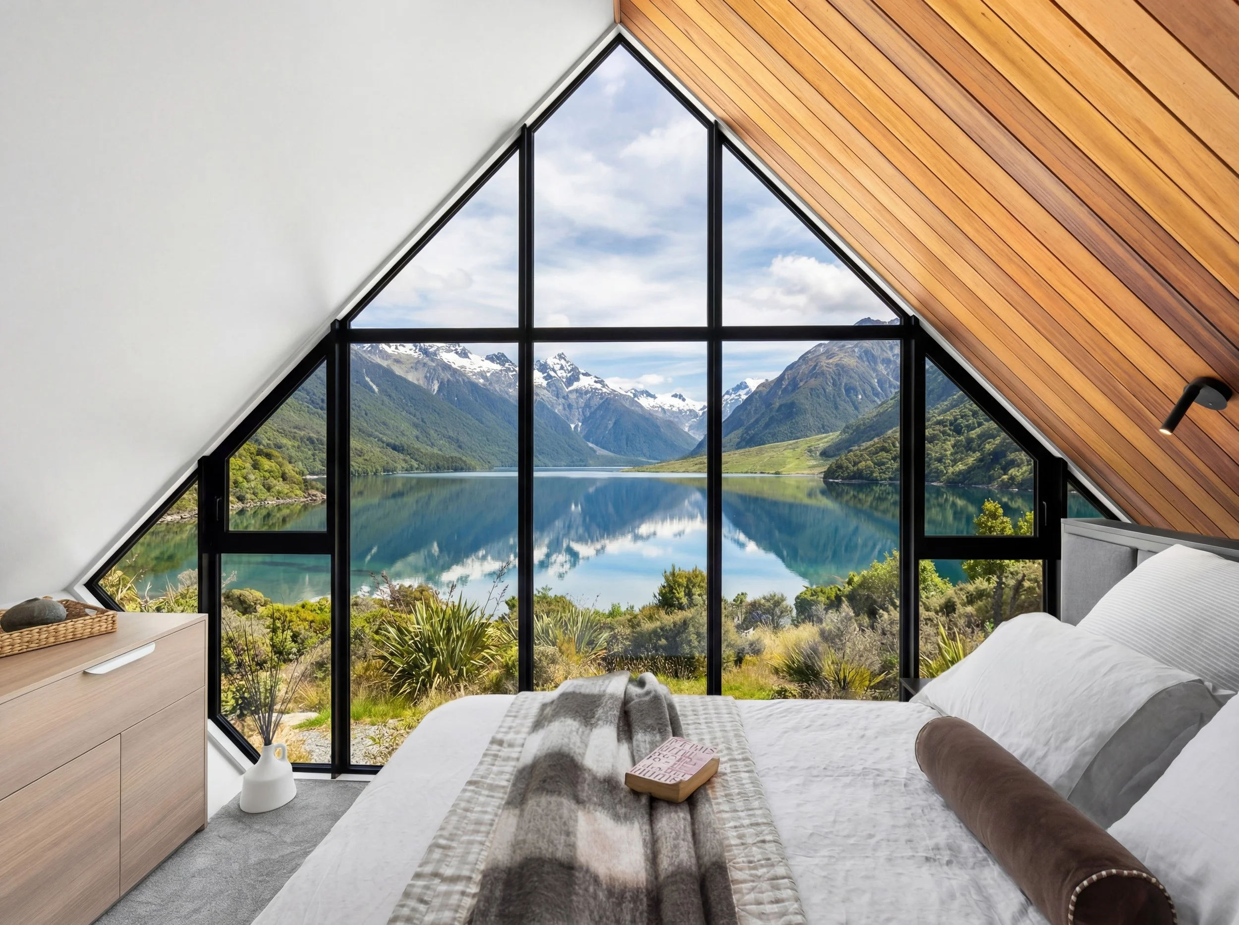 Interior of a bedroom with a large window overlooking a mountain lake in a scenic landscape.