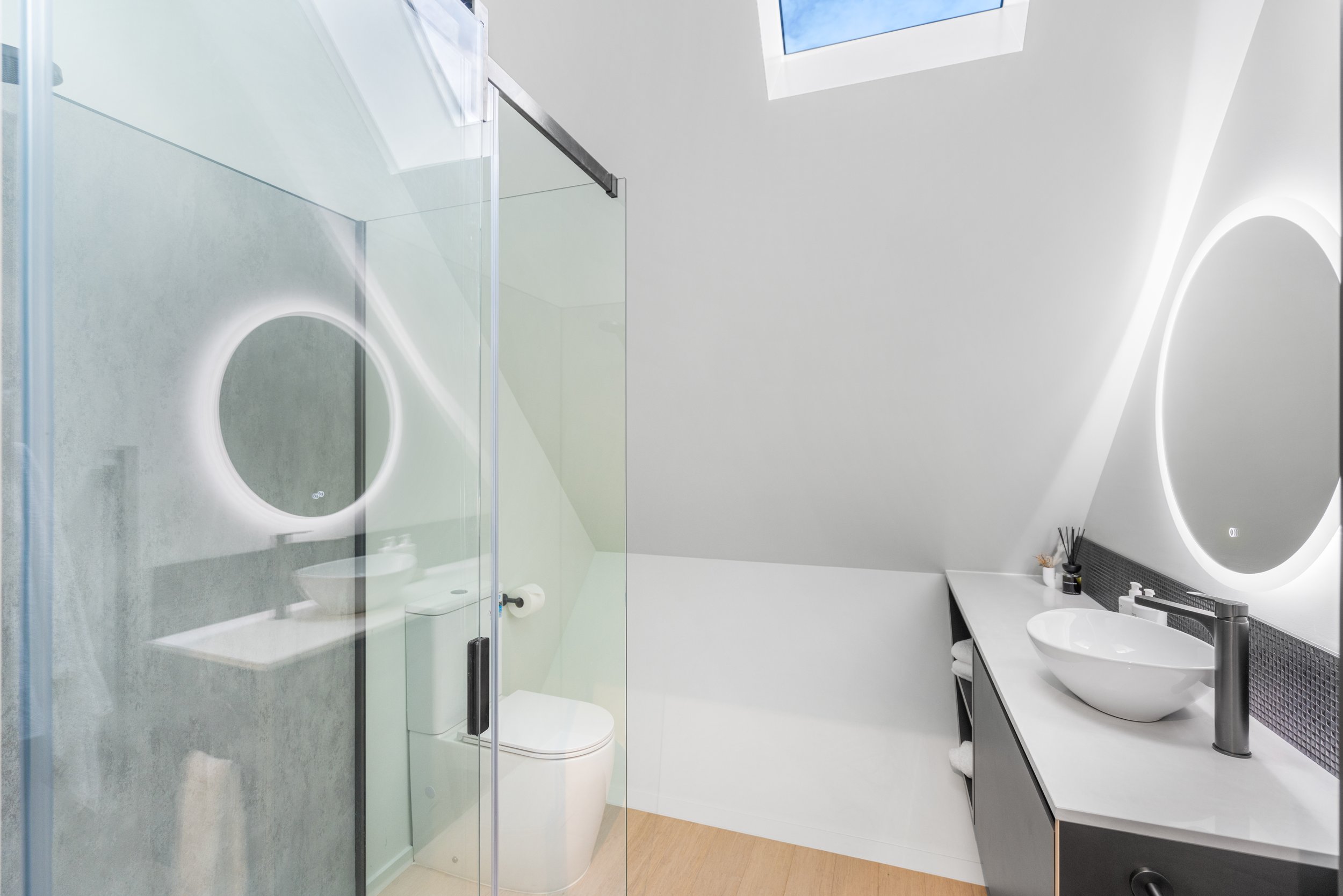 Modern bathroom with white sink and mirror with backlight, toilet, and glass-enclosed shower.