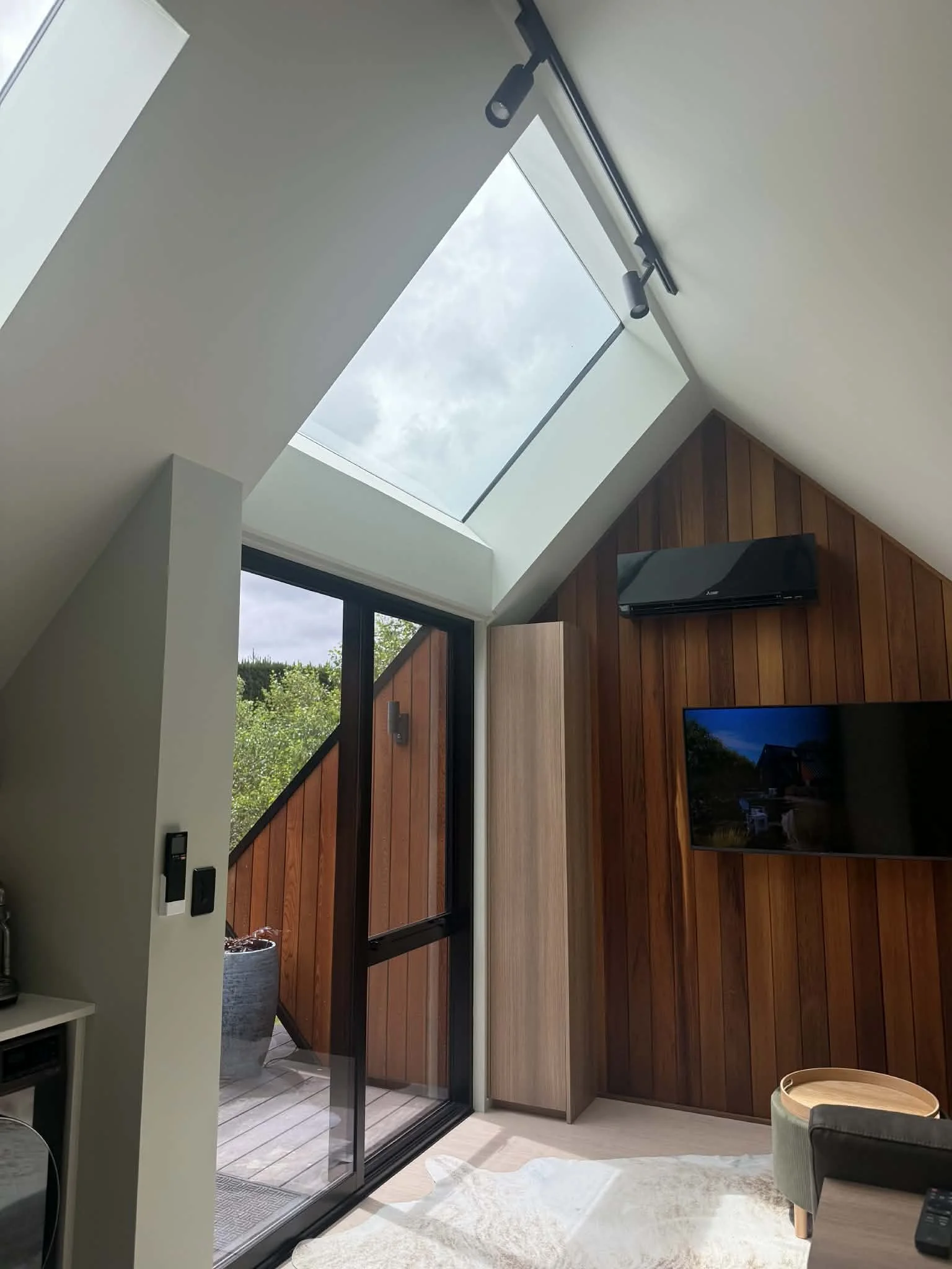 Modern living room with a large skylight, a wall-mounted TV, and a glass door leading to a small balcony with outdoor plants.