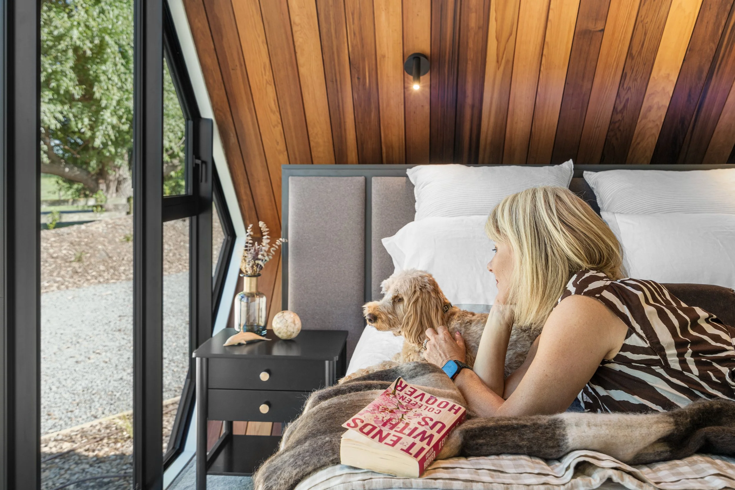 A woman lying in bed with a dog, looking out the window. The room has wood-paneled walls, a black nightstand with decorative items, and a book titled 'Famous with Out H