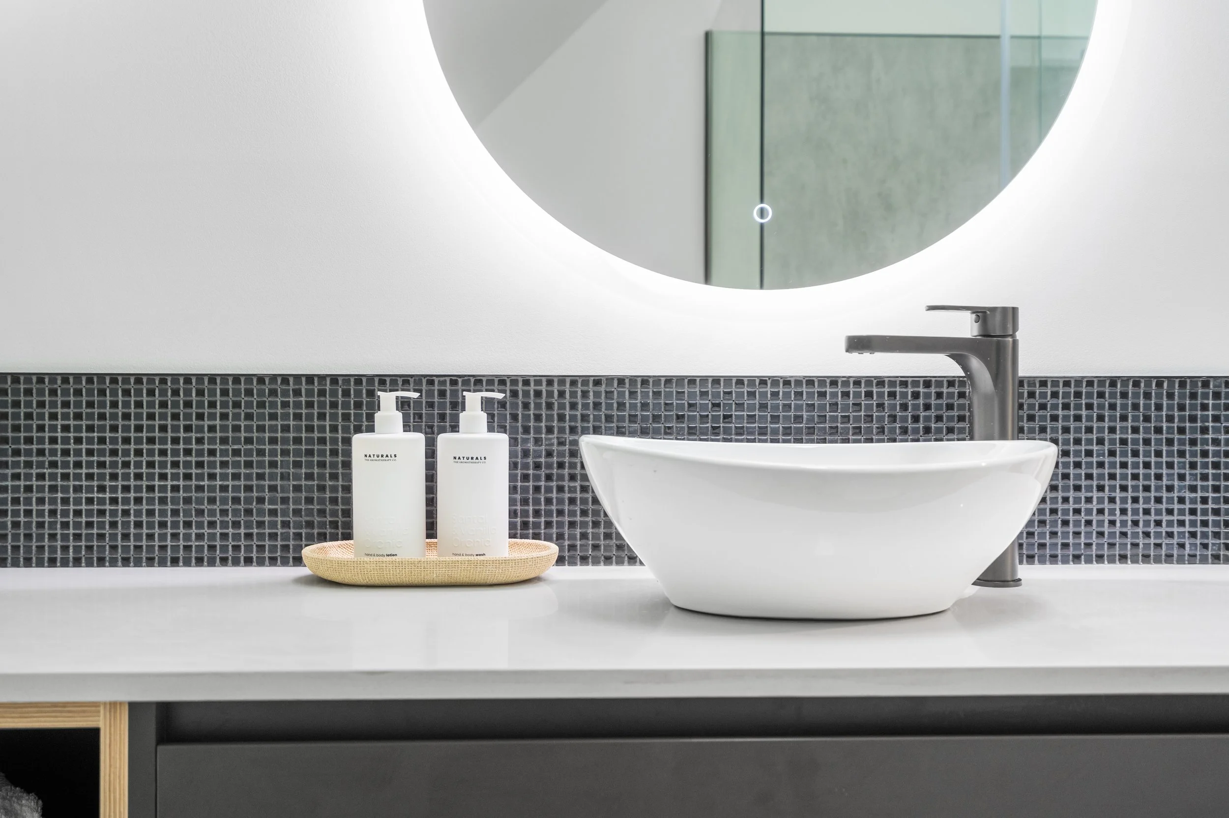 Bathroom sink area with a white vessel sink, a black faucet, a round mirror, and two white soap dispensers on a beige tray.