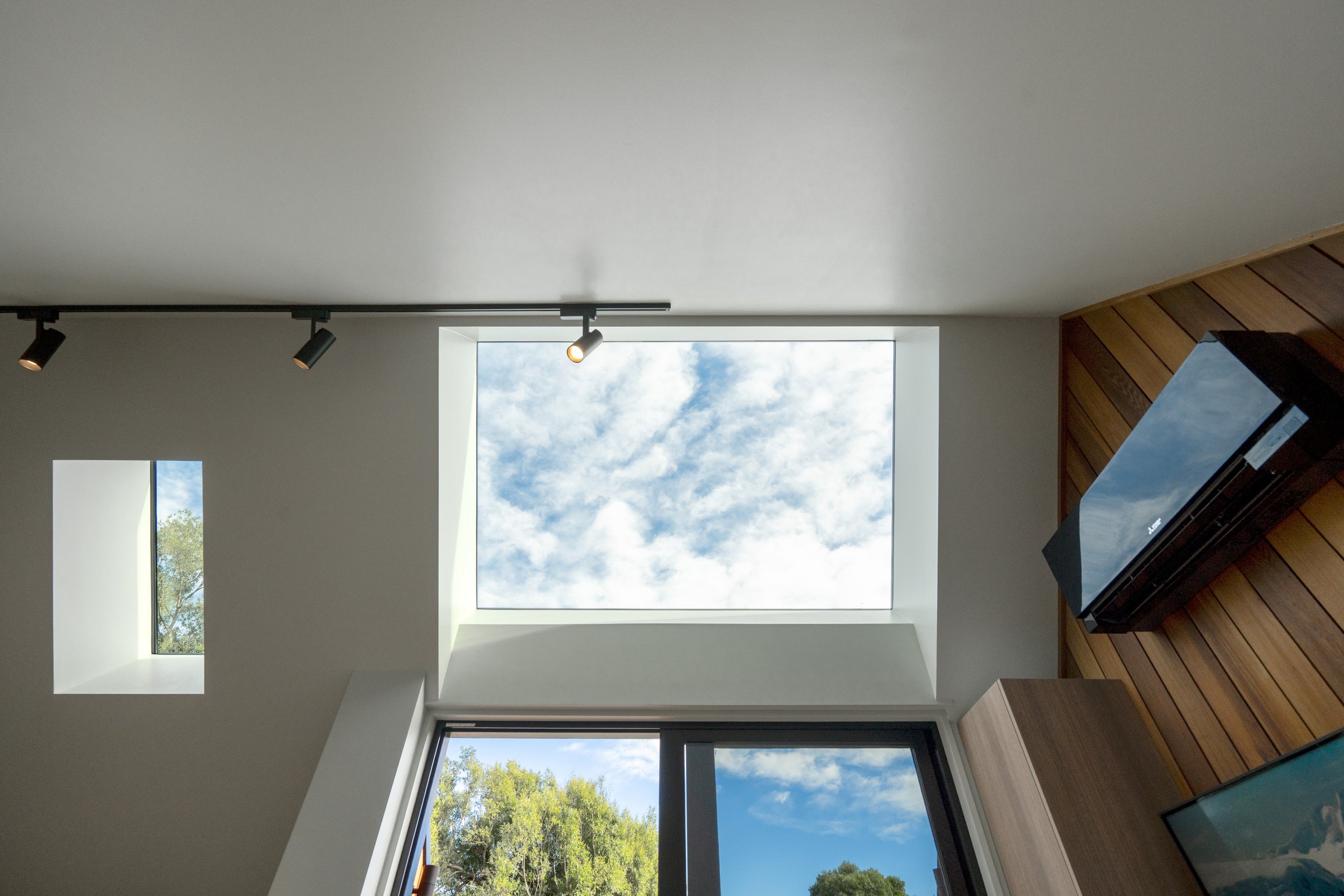 Interior view of a room with a large skylight showing a blue sky and clouds, a smaller window with trees outside, and a television mounted on a wooden wall.