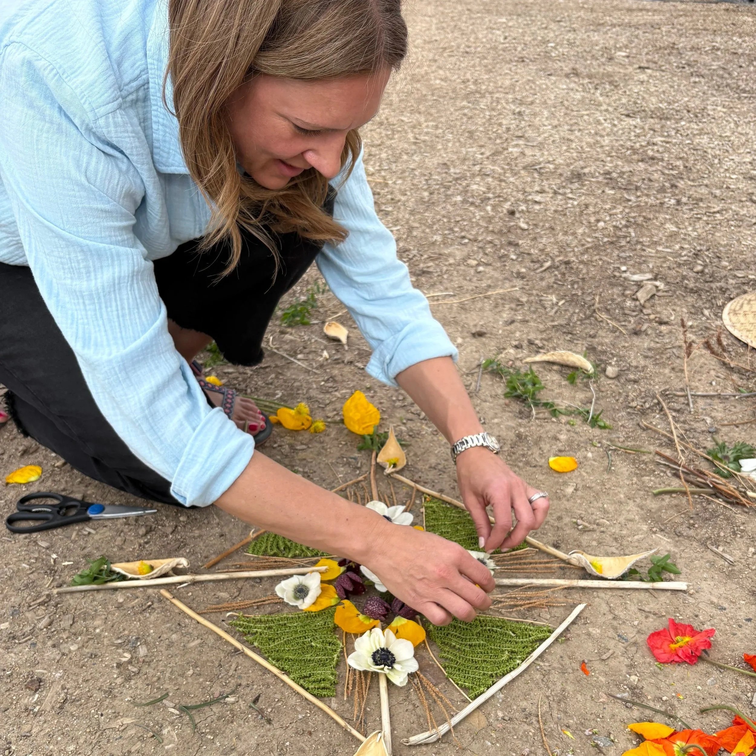 A woman arranging flowers on the ground, creating a floral pattern with various colorful flowers and green foliage on a dirt surface.