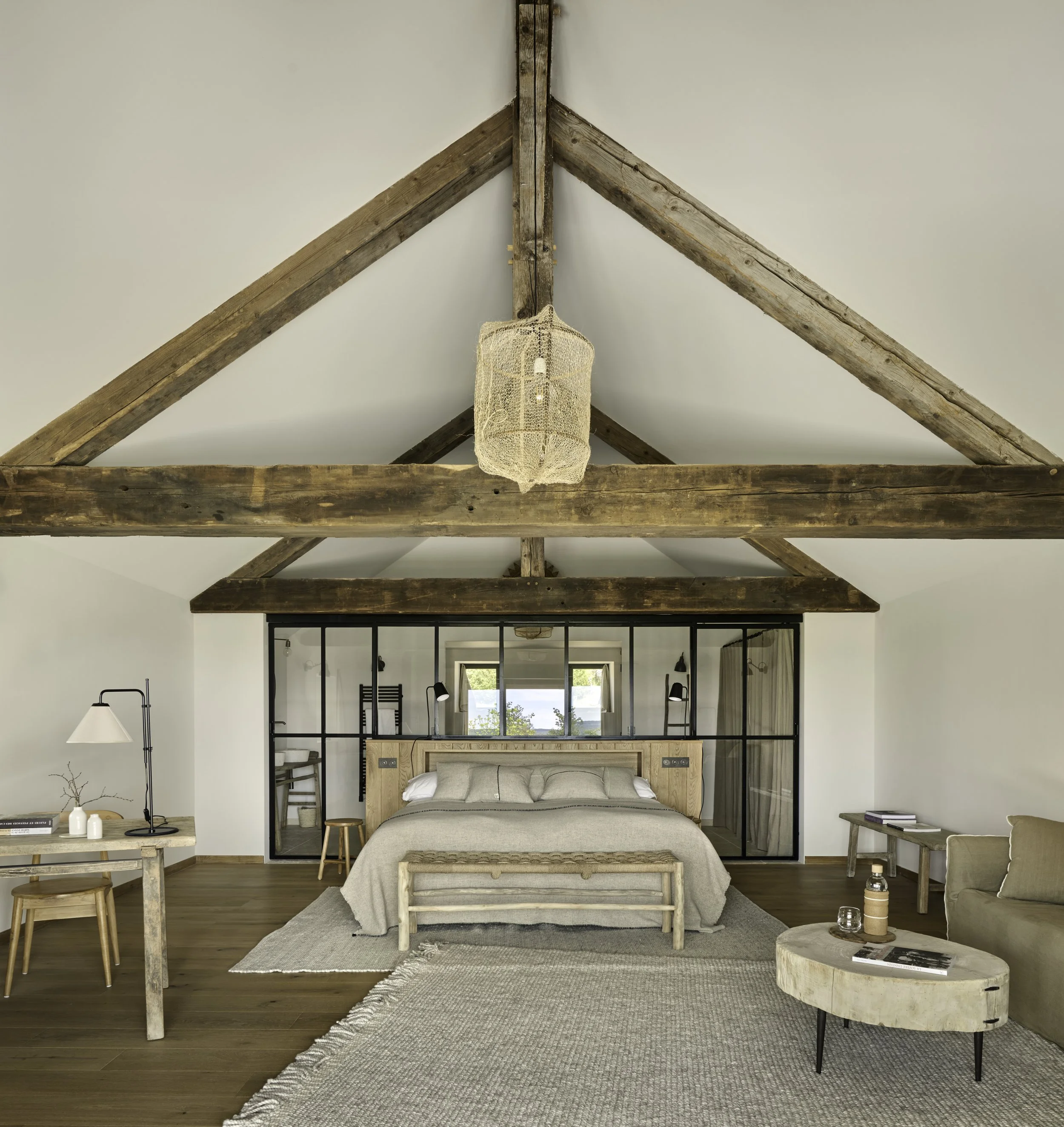 Modern bedroom with white walls, wooden floor, and vaulted ceiling with exposed wooden beams. Features a bed with neutral bedding, a hanging wicker light, a rustic wooden bench, a round side table with a candle and mug, a beige sofa, and a workspace with a wooden table, chairs, and a desk lamp. Large window behind the bed shows outdoor greenery.