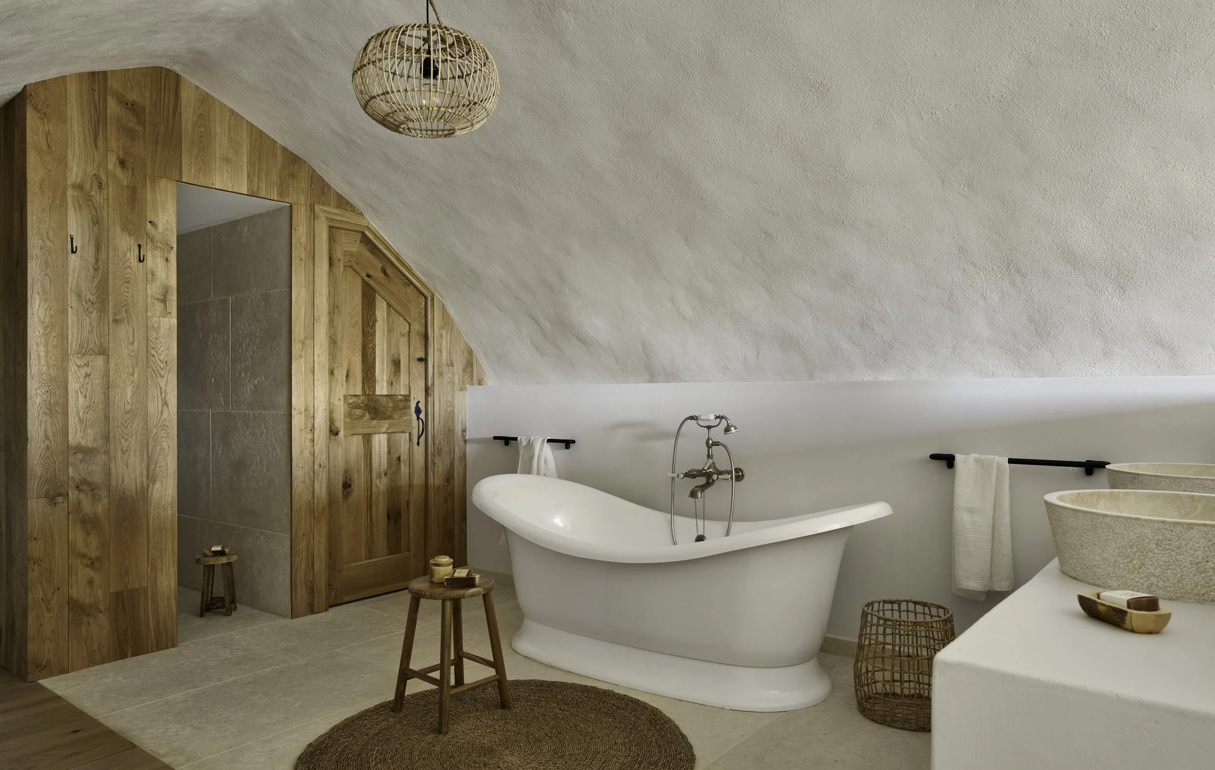 A bathroom with a freestanding clawfoot tub, a round wooden stool, stone sinks on the counter, and a wooden door and wall paneling with a sloped ceiling.
