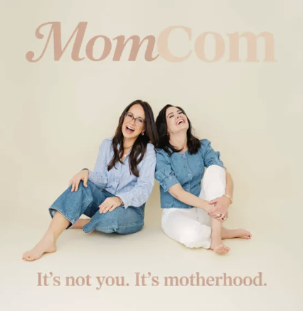 Two women sitting on the floor, smiling and laughing against a light background with a 'MomCom' logo and the phrase 'It's not you. It's motherhood.'