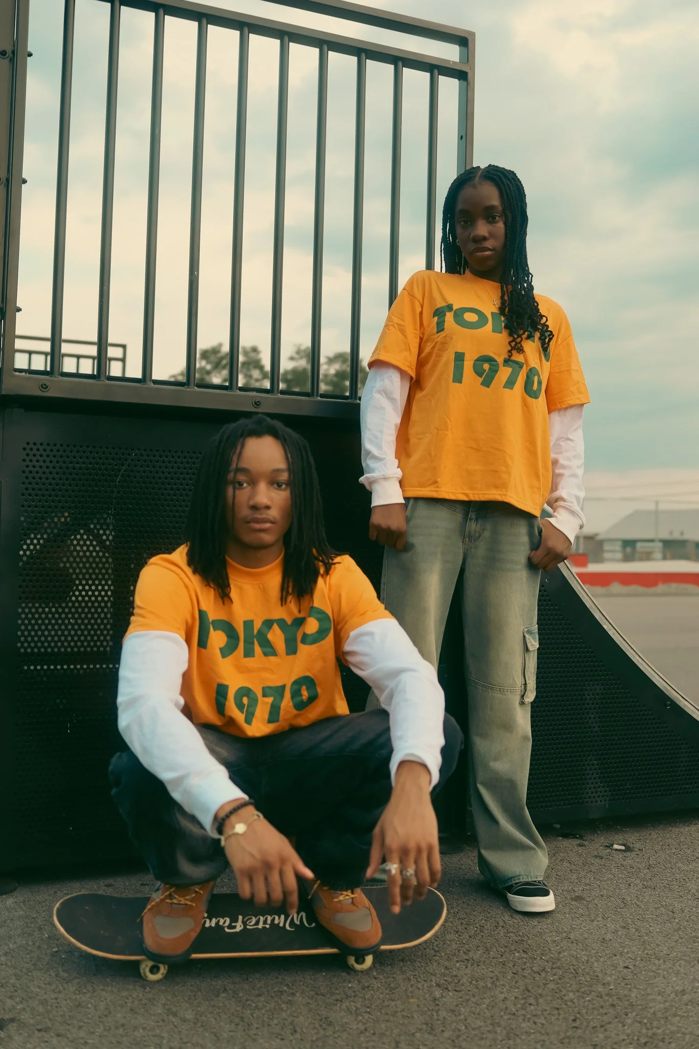 Two young women with dreadlocks wearing matching yellow T-shirts with green text, one squatting on a skateboard and the other standing in an outdoor skate park, against a cloudy sky.