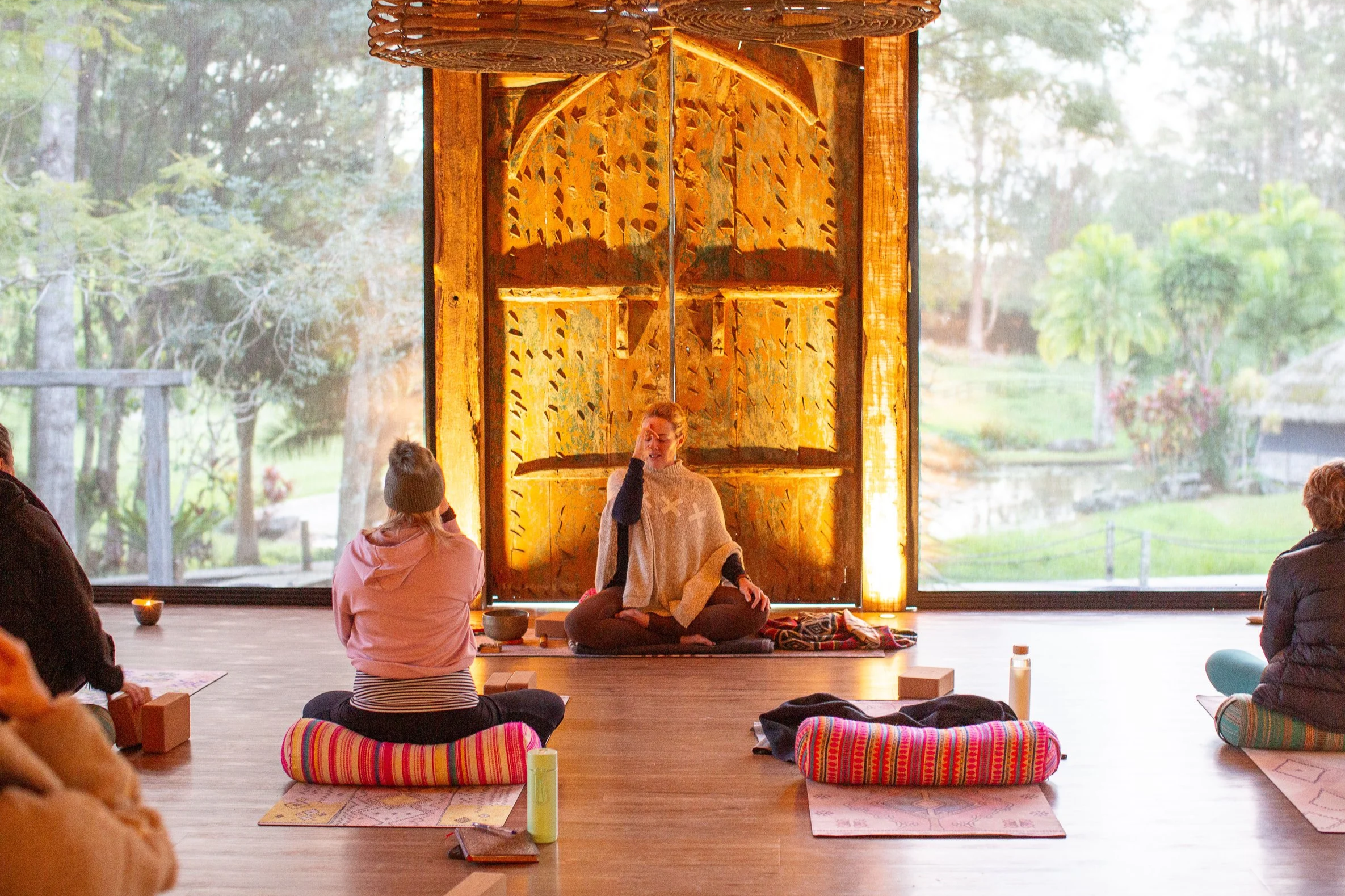 A woman leading a meditation or yoga session in a spacious room with large windows showing trees and greenery outside. She is seated on a cushion and gesturing with her hand, with others sitting on mats and cushions around her.