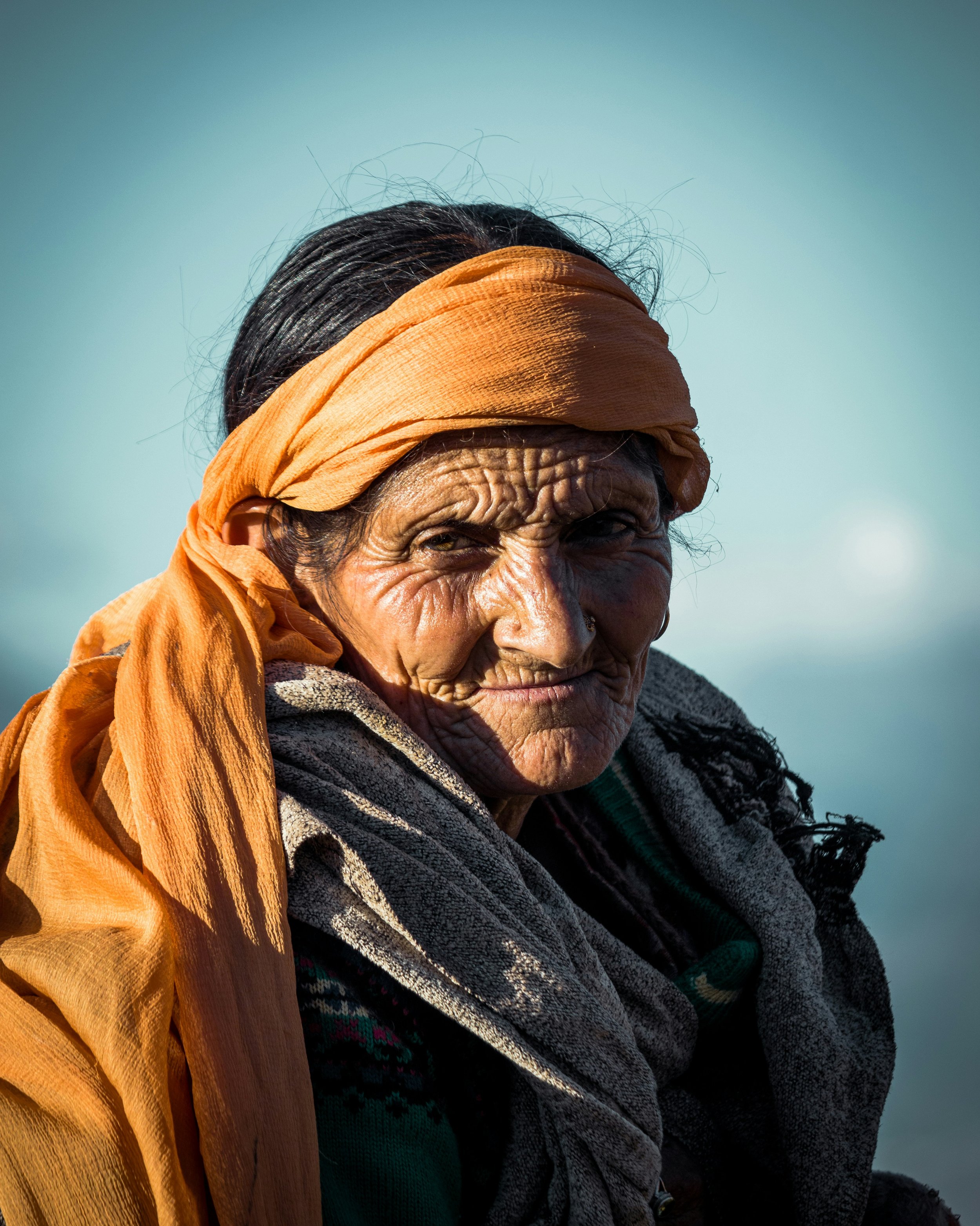 Close-up of an elderly woman with deep wrinkles, wearing an orange headscarf and gray clothing, looking directly at the camera against a clear sky background.