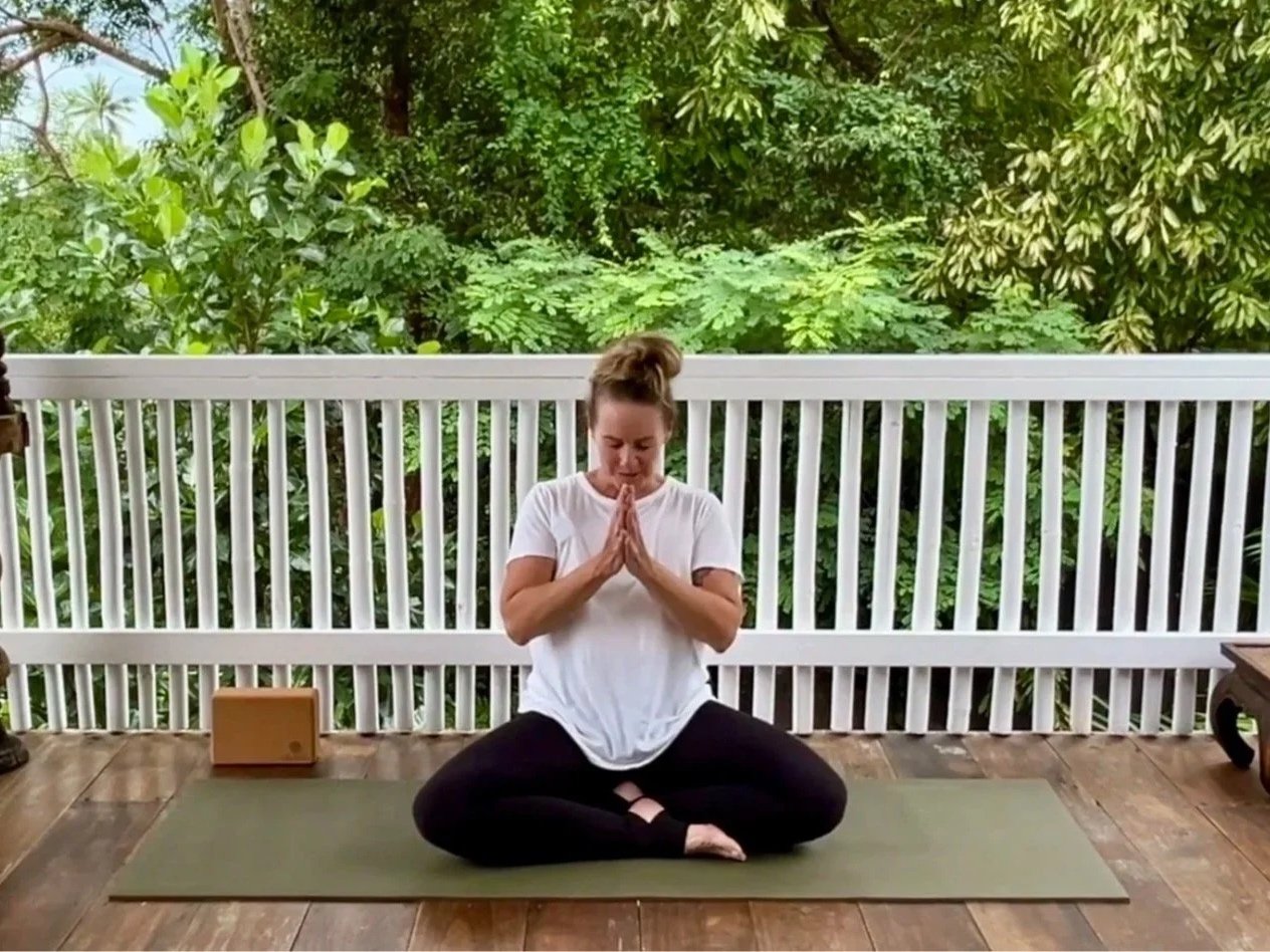Woman practicing yoga or meditation on a balcony with lush green trees in the background.