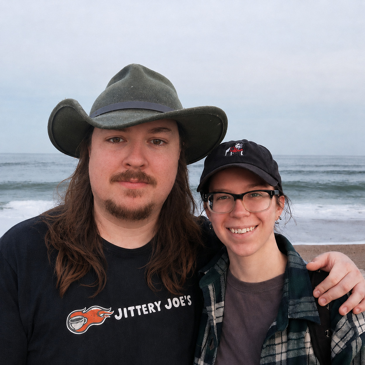 A man and woman taking a selfie on a beach with the ocean in the background. The man is wearing a wide-brimmed hat and a black T-shirt. The woman is wearing glasses, a black cap, and a plaid shirt.