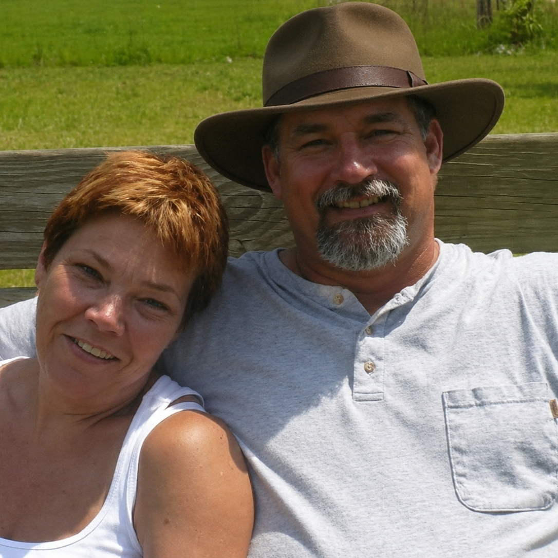 A smiling woman with short red hair and a bearded man wearing a wide-brimmed hat, sitting close together outdoors on a wooden bench in a grassy area.
