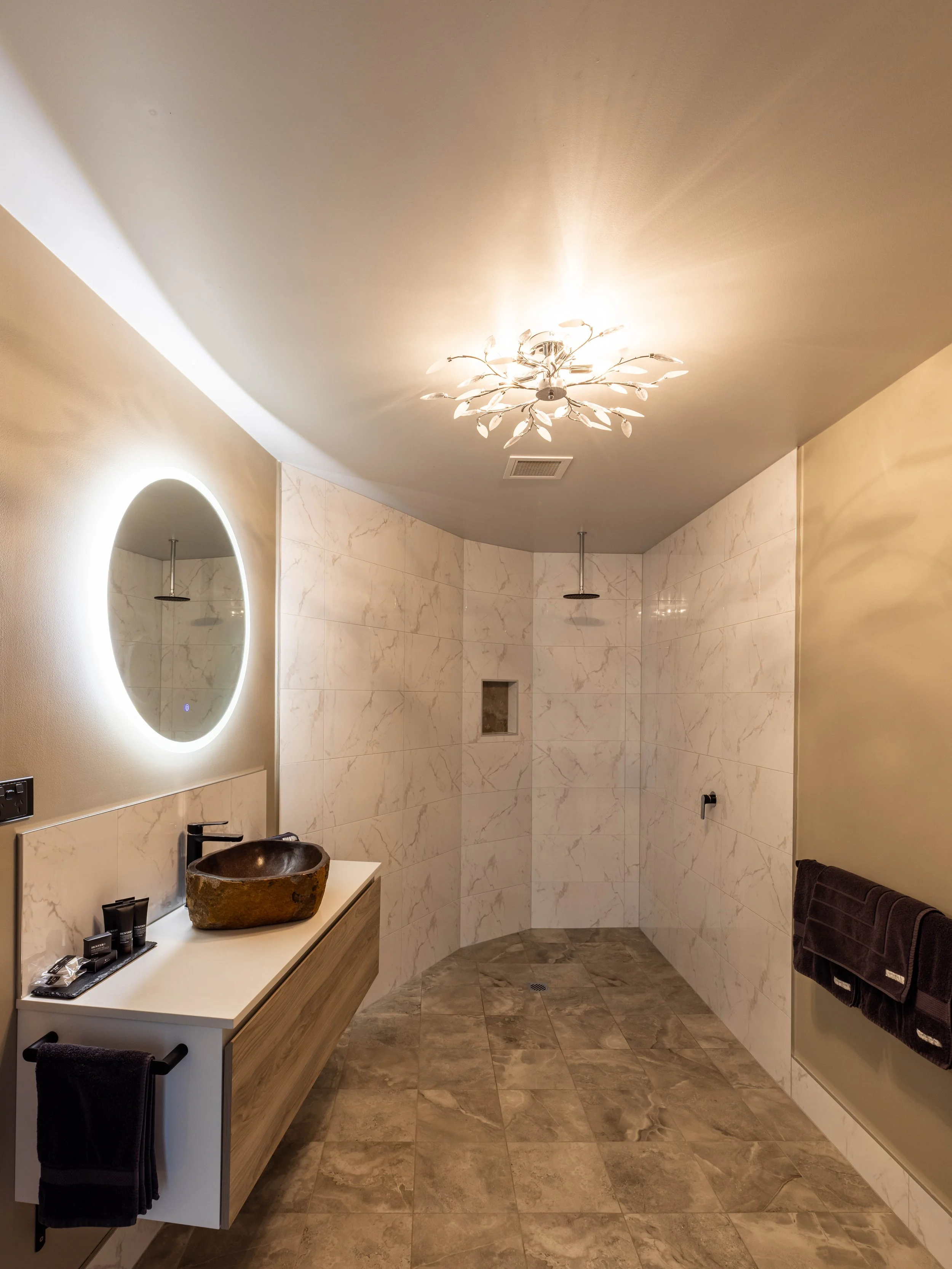 Modern bathroom with marble walls, circular mirror, unique stone sink, and decorative ceiling light.