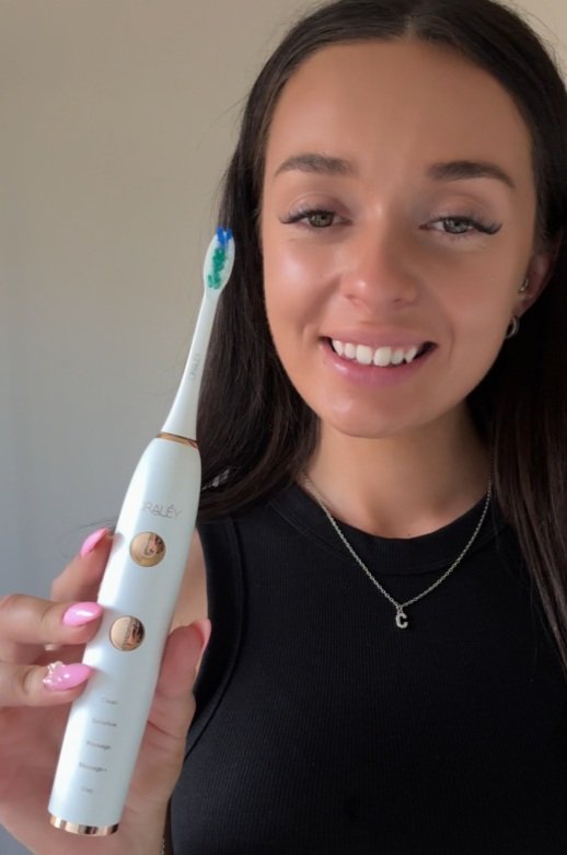 A young woman with dark hair smiling and holding a white Oraley electric toothbrush with blue and green bristles, wearing a black top and a silver necklace with a letter 'C' pendant.