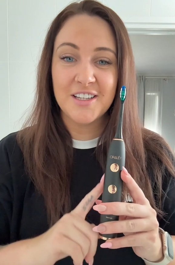 Woman with long brown hair holding an Oraley electric toothbrush and pointing at it.