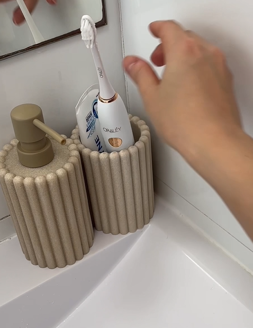 A hand reaching for an electric toothbrush in a decorative beige holder next to a matching soap dispenser on a bathroom countertop.