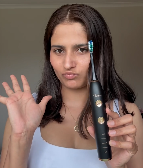 Woman with dark hair holding a black Oraley electric toothbrush, making a playful face with one eye slightly squinted and her lips puckered, while waving with her other hand.