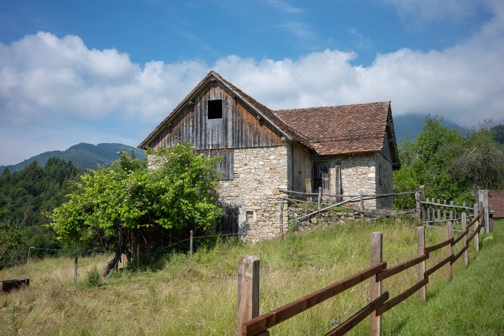 Old stone and timber stavolo with a weathered wooden gable, an empty dark window, and a rough wooden fence in the foreground