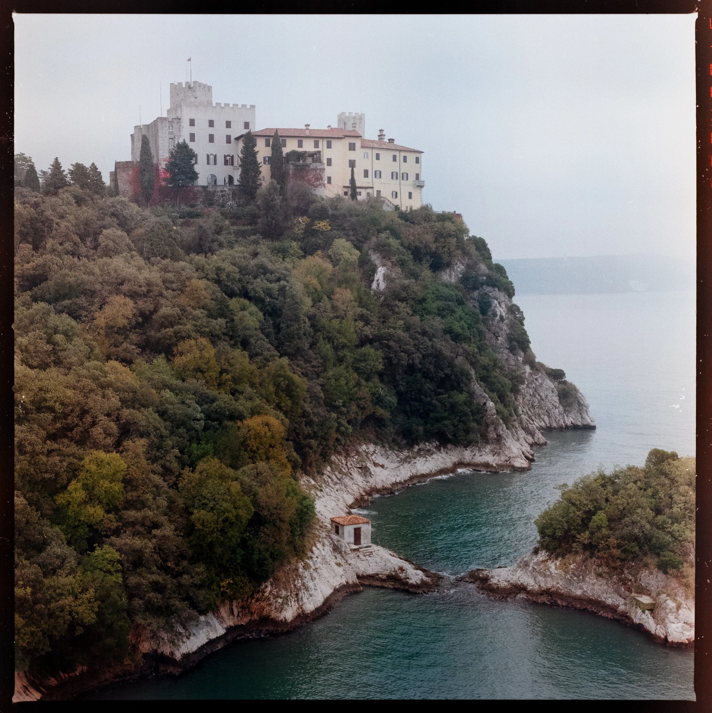 View of Duino Castle on a cliff above the Adriatic Sea, seen from the ruins of the old castle, with a small bay and stone hut below surrounded by greenery