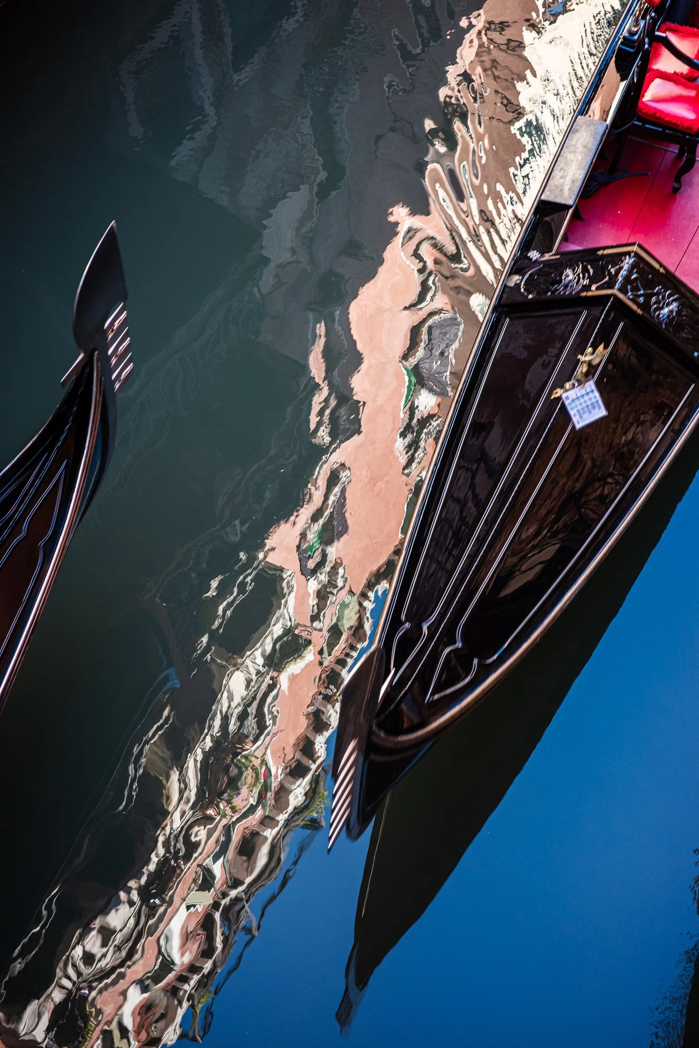 Two black gondolas at the edges of a narrow Venetian canal, the dark water between them reflecting the colorful buildings above as rippled, abstract bands of peach, white and green.