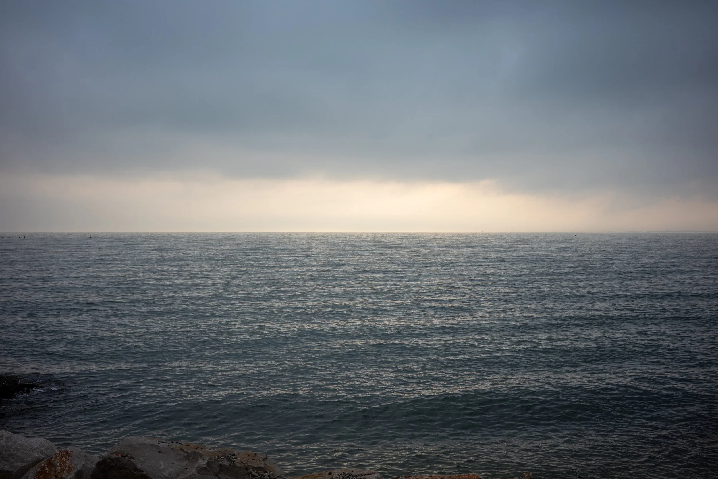 View of the Adriatic Sea at dusk with soft light breaking through the clouds, seen from the rocky shore near the end of the Rilke Trail