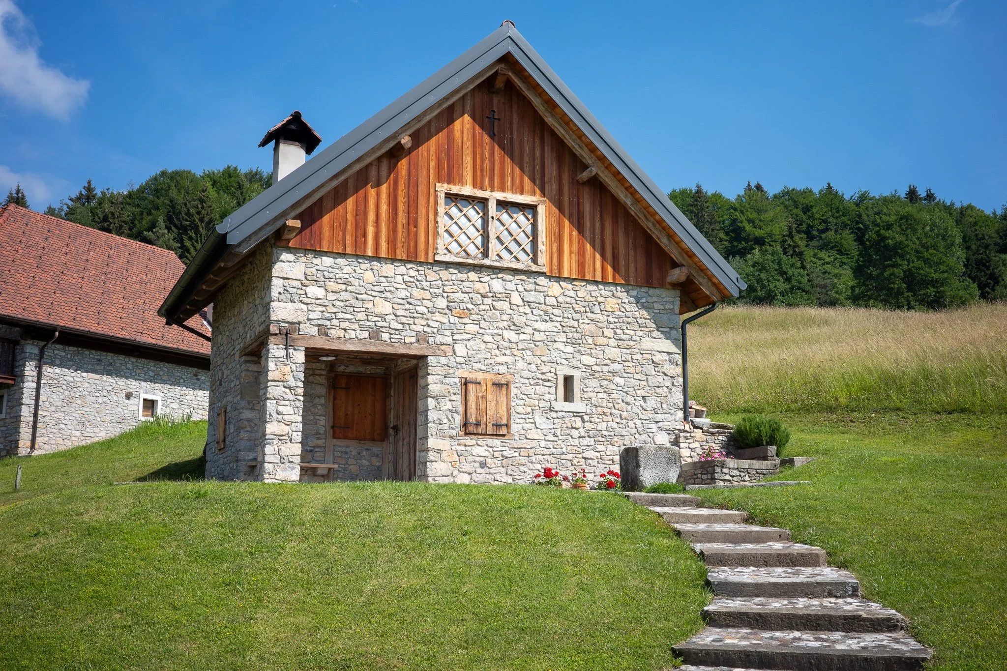 Restored stone and timber stavolo with a lattice window and stone steps climbing the grassy slope beside it