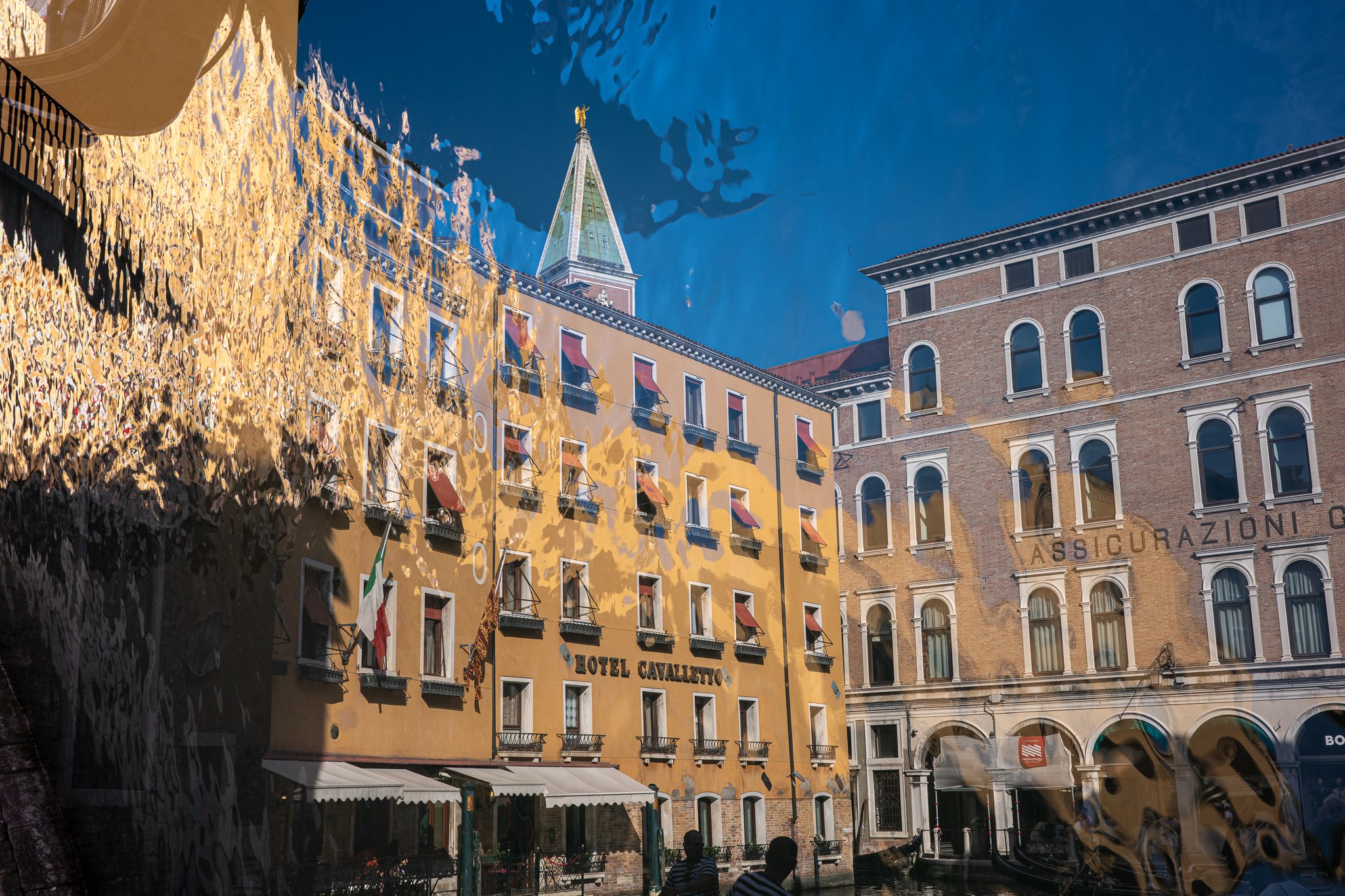 A double exposure of the Hotel Cavalletto with the campanile of San Marco behind it, rippled water overlaid across the sky so the blue takes on a watery, dappled texture, and golden reflections washing over the left of the frame.