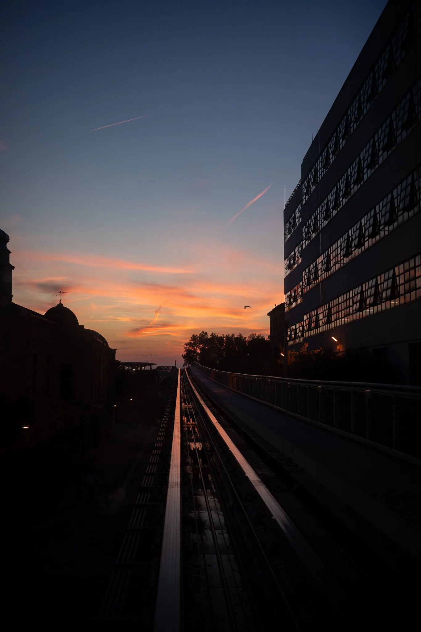 The twin tracks of the Venice People Mover receding toward a distant vanishing point at dusk, with an orange-and-blue sunset sky above, a lit building to the right and a church dome silhouetted to the left