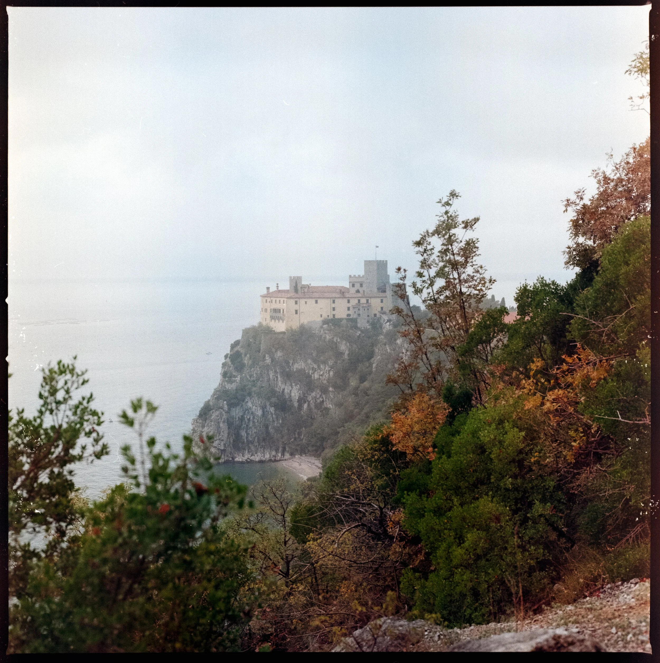 View of Duino Castle perched on a cliff above the Adriatic Sea, partially framed by trees along the Rilke Trail on an overcast day