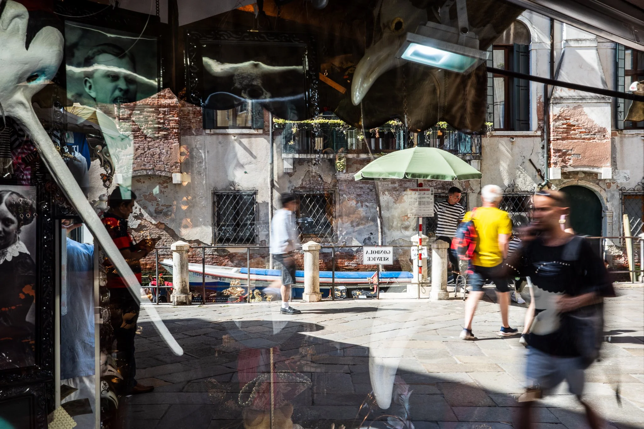 Venetian carnival masks, including a long-beaked plague-doctor mask, displayed in a shop window that reflects the sunlit street behind — people walking past weathered brick buildings, with a reversed "Gondola Service" sign showing through the glass.