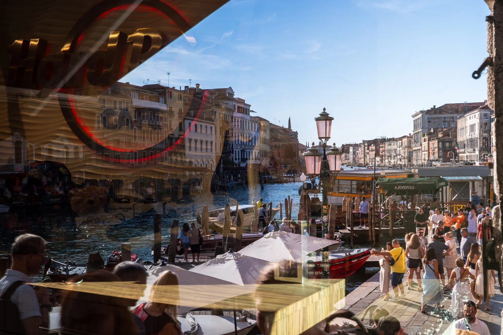 The Grand Canal near the Rialto viewed through a café window, with a reflected gold "Hard Rock" logo overlaying the scene — crowds along the embankment, moored boats, an ornate triple lamp post and a "Gondola" sign by the water.