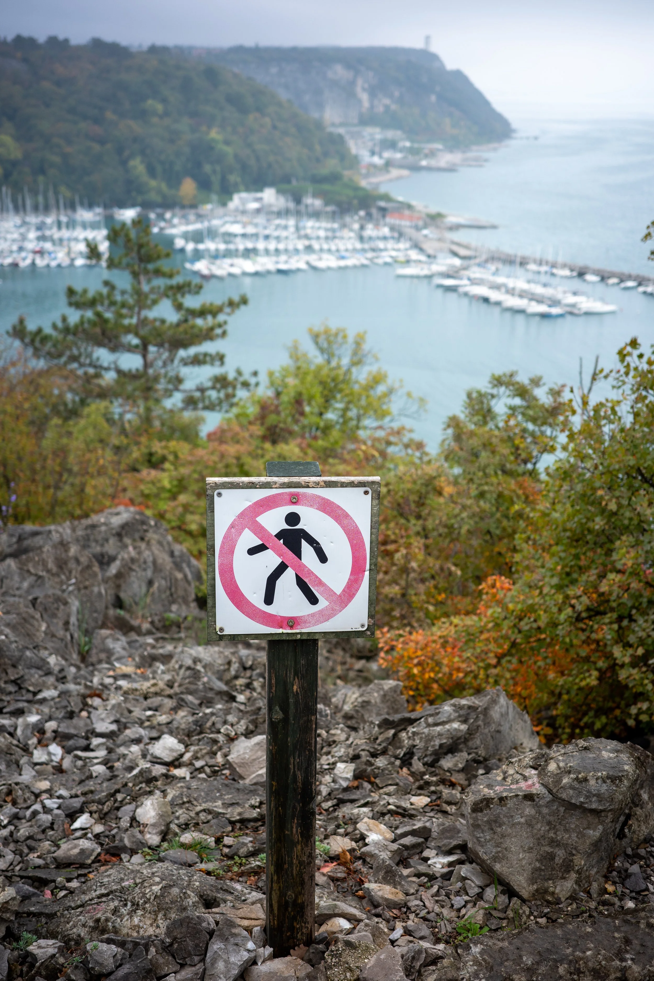 A warning sign along the Rilke Trail near Trieste, placed on a rocky cliff above a marina filled with boats and surrounded by autumn trees