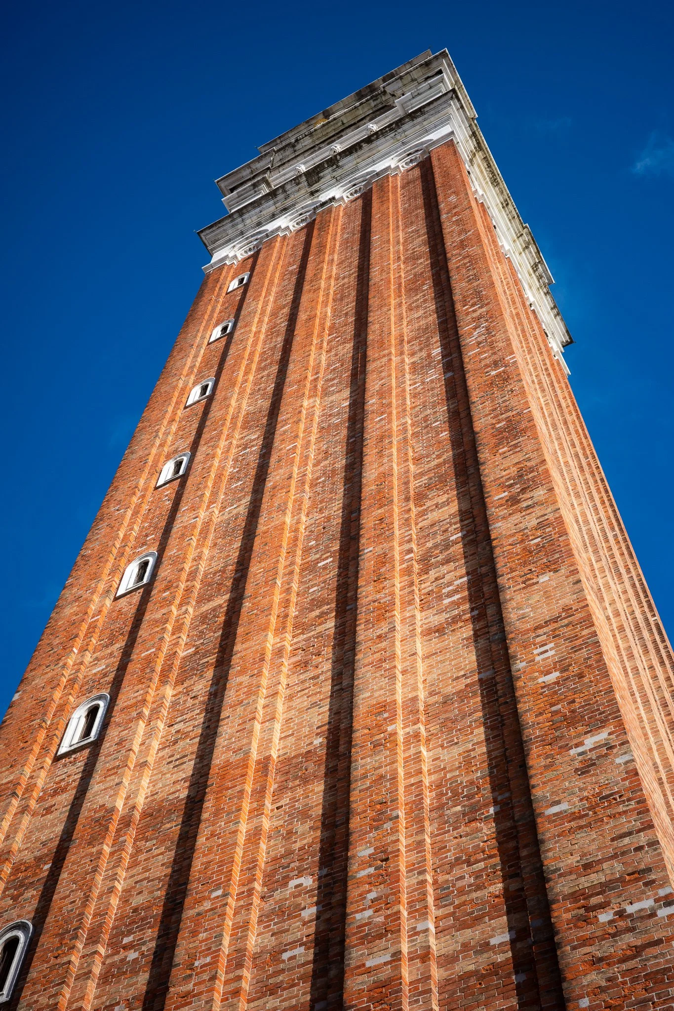 The red-brick shaft of St Mark's Campanile seen from its base, its converging lines rising toward the white stone belfry against a deep blue sky.