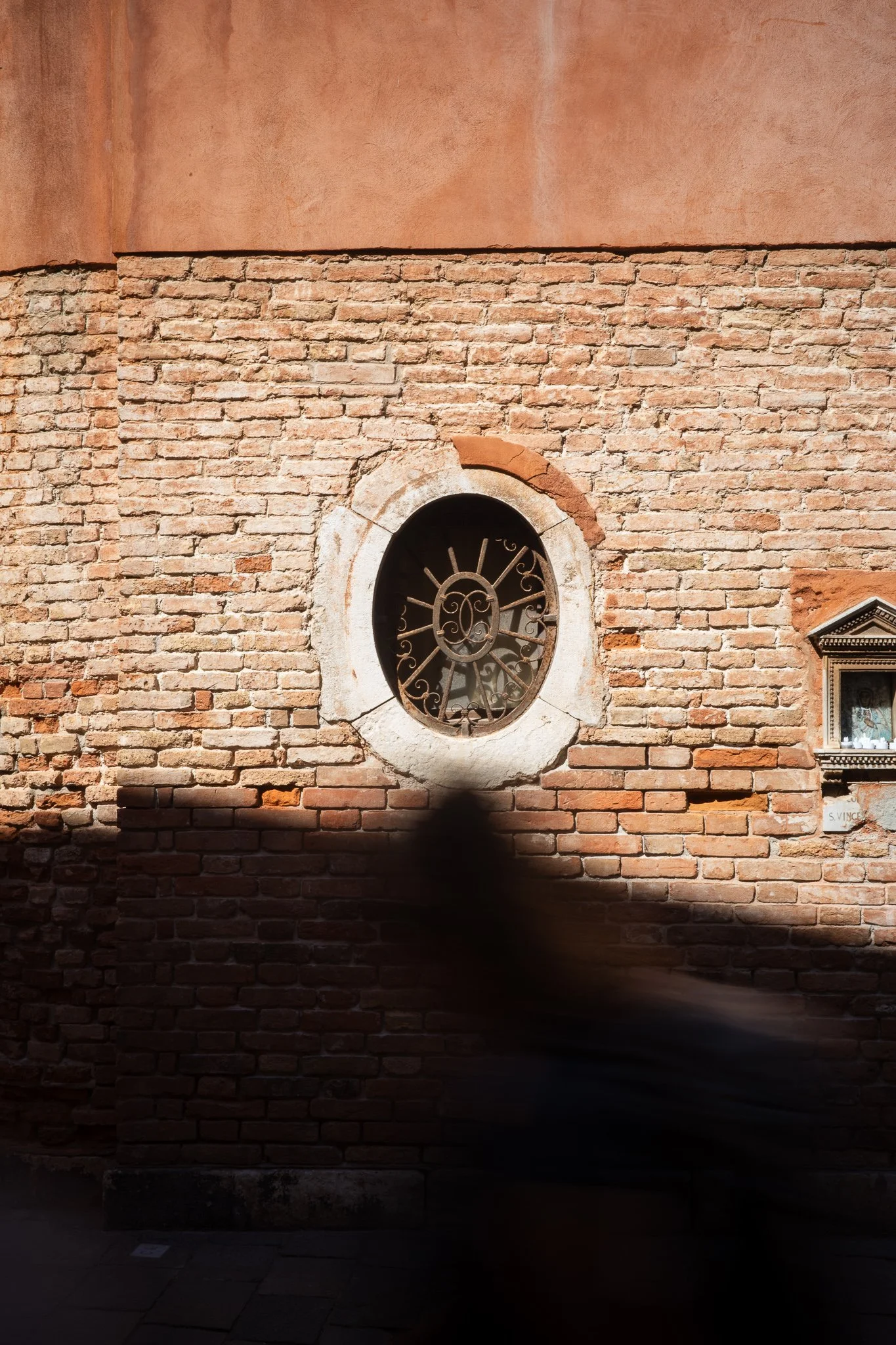 Oval iron-grilled window set in a sunlit Venetian brick wall, with a small votive shrine at the right edge and the motion-blurred figure of a person walking past in the foreground.