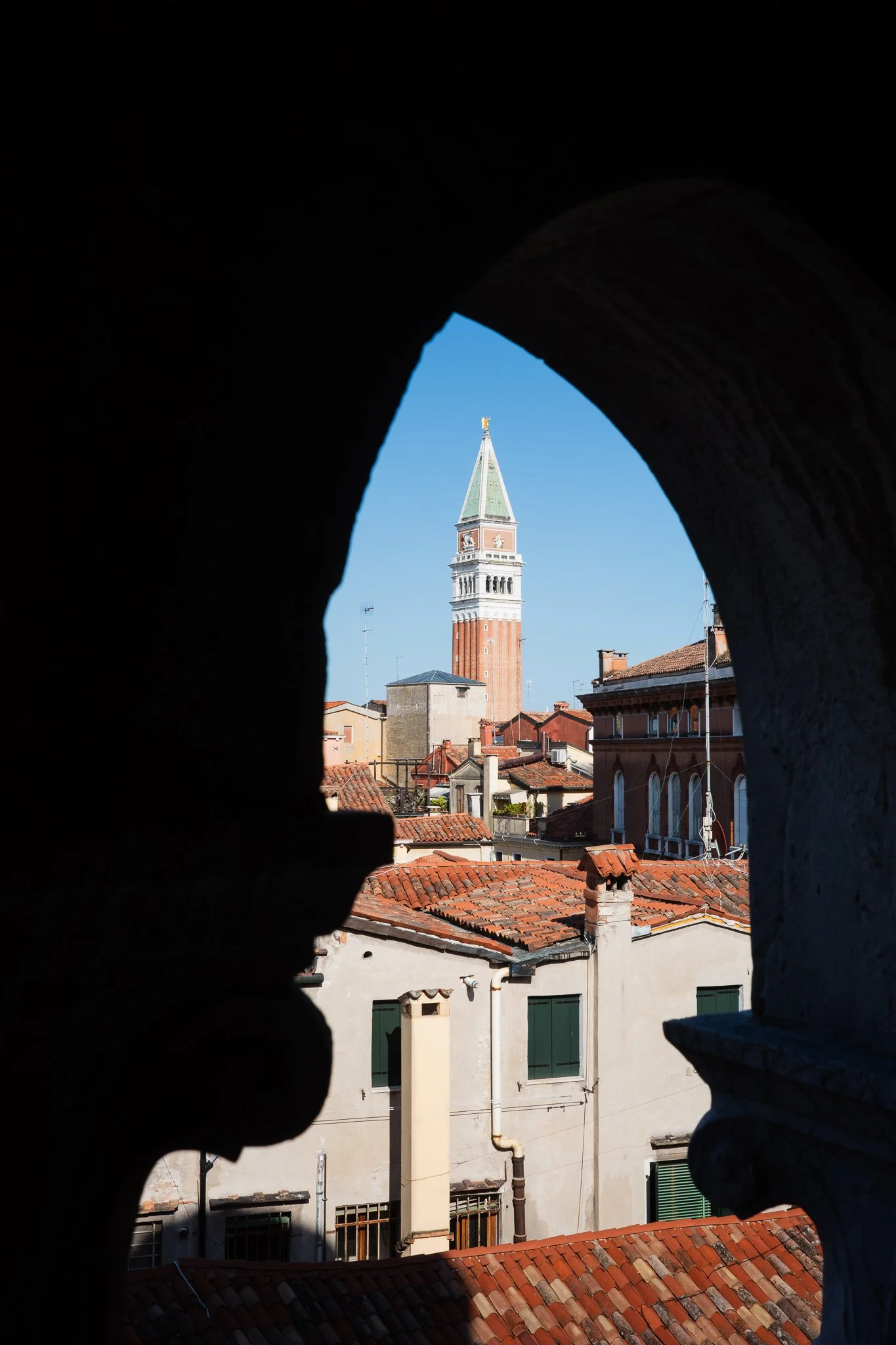 The campanile of San Marco rising above Venetian tiled rooftops, framed by the silhouetted arch of the Scala Contarini del Bovolo against a clear blue sky