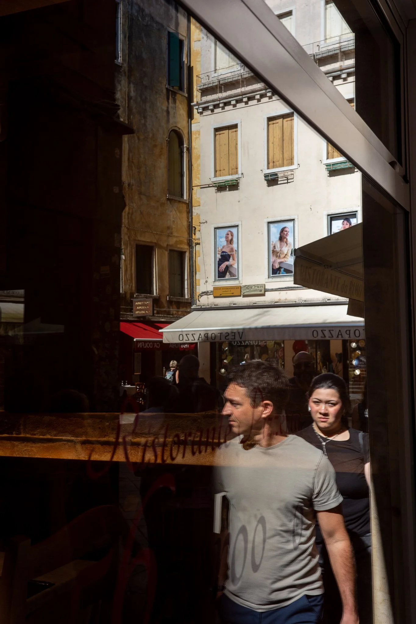 A man and woman reflected in a Venetian restaurant window as they walk past, layered over the sunlit calle behind — shuttered buildings, a café awning, and fashion posters with models showing through the glass.