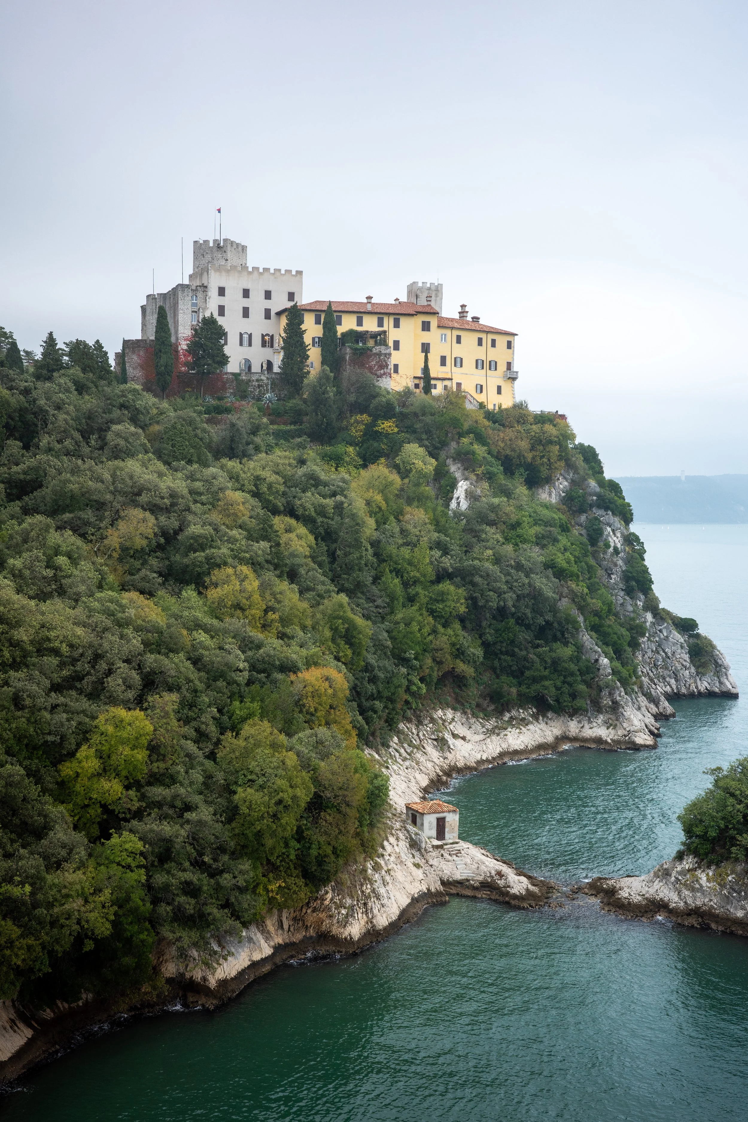 View of Duino Castle perched on a cliff above the Adriatic Sea, with a small bay and stone hut below, surrounded by dense green trees on an overcast day
