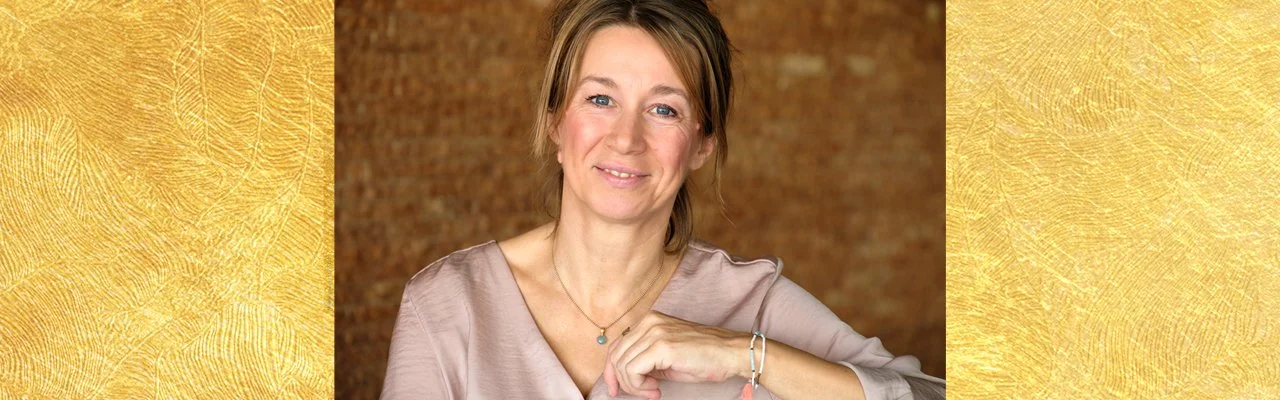 Ein Lächelnde Frau mit braunem Haar, trägt eine silberne Halskette und Armbänder, sitzt vor einem braunen, modernen Hintergrund.