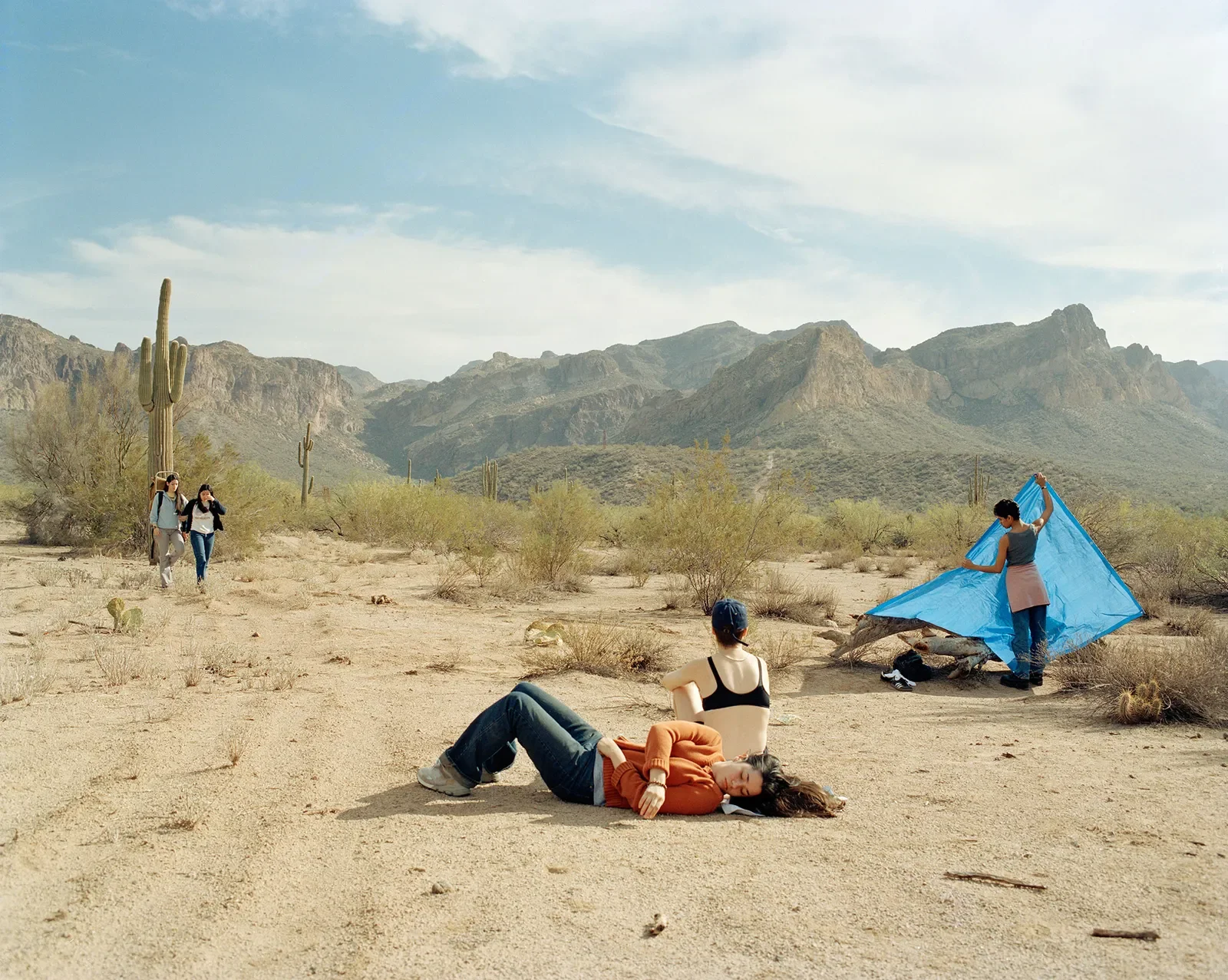 Three women relaxing on the desert ground, one lying down, two sitting, while two others set up a blue tent in a desert landscape with cacti and mountains in the background.